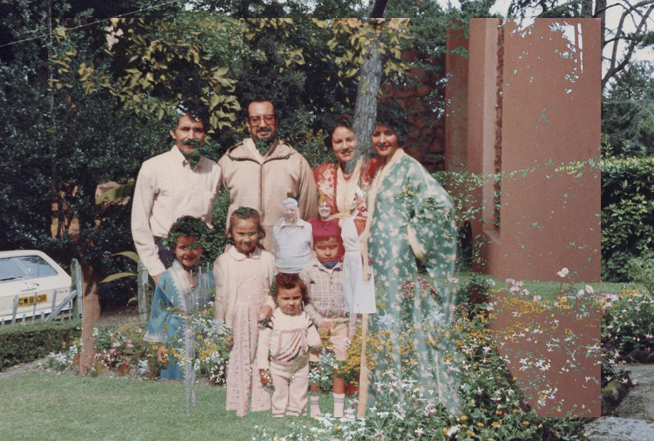 A family group photo outdoors with three adult men, two adult women, and four young children, surrounded by trees, bushes, and flowers.