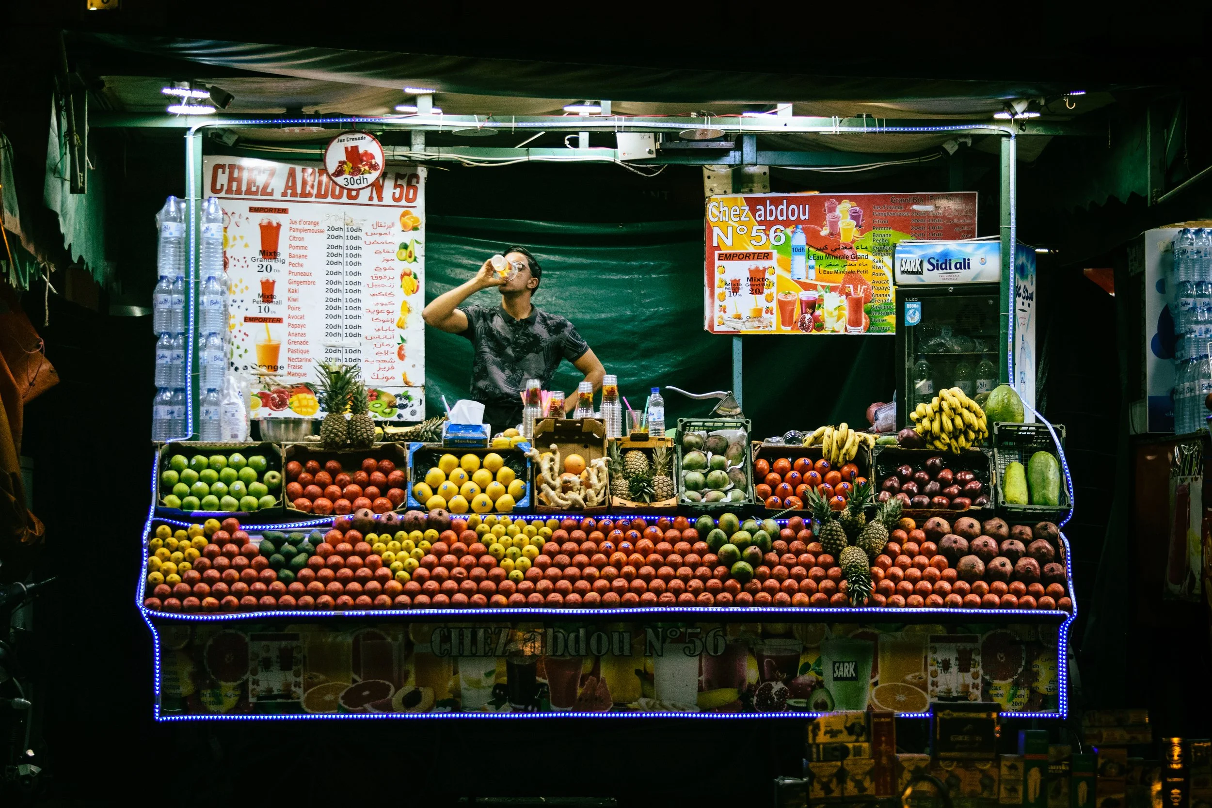 Night market fruit stand with an array of fresh fruits including apples, pineapples, bananas, watermelons, and pomegranates. A man is drinking from a plastic cup behind the stand. Colorful signs display menu items and prices, with bottled water on di
