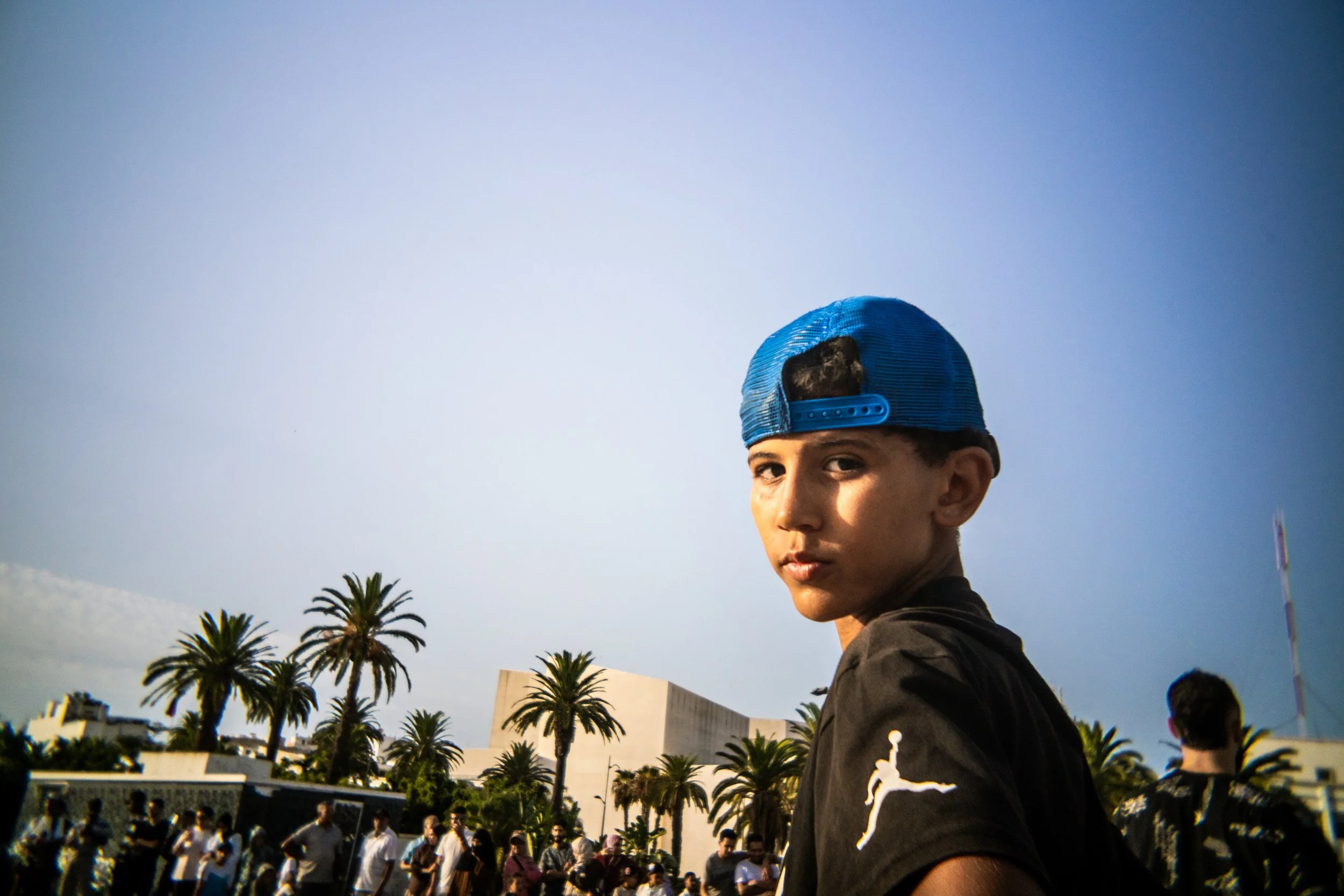 Young man wearing a blue backward baseball cap and black Jordan shirt outdoors with palm trees and a crowd