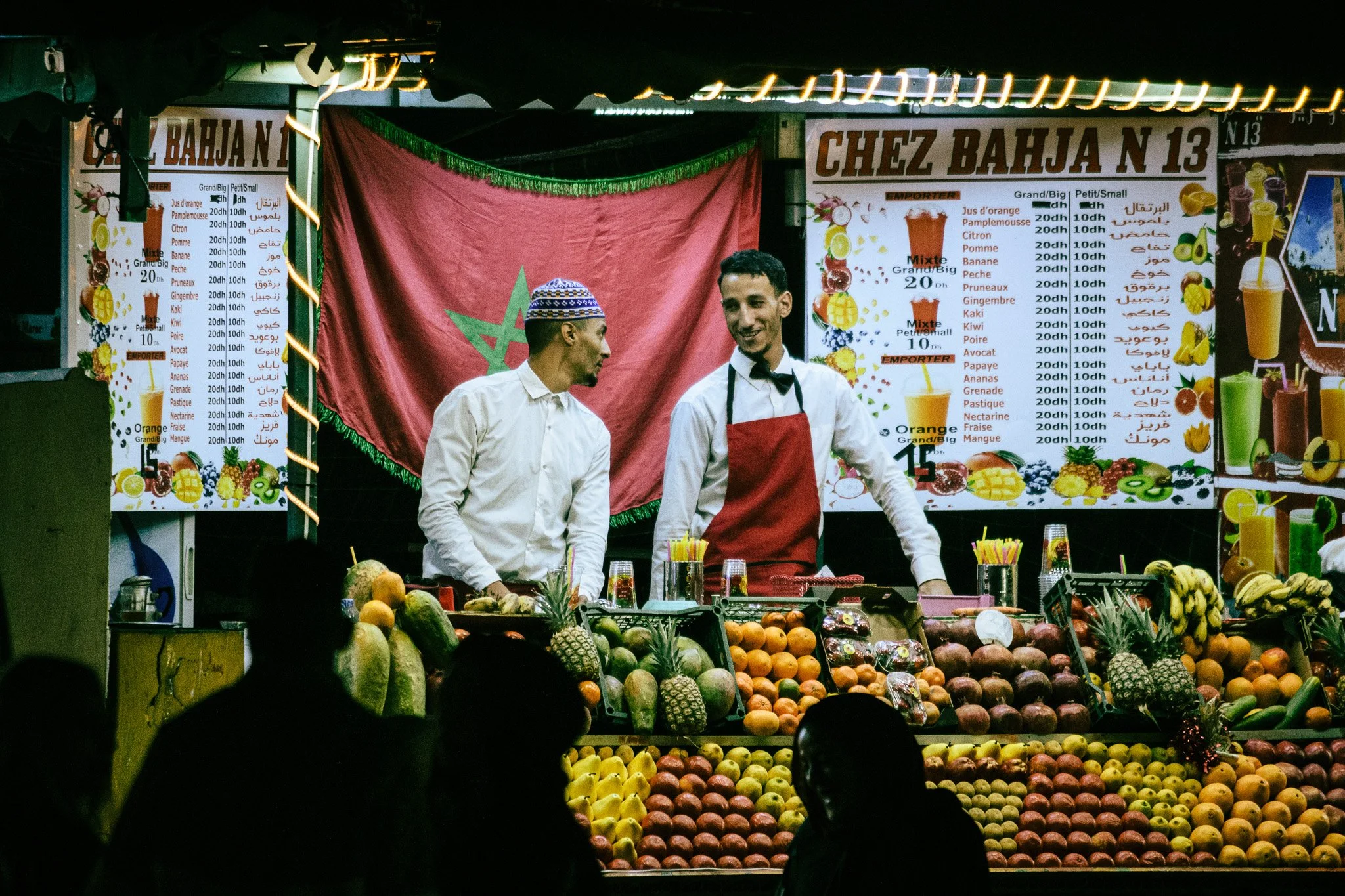 A fruit stand at night with two men in white shirts and black bow ties, one wearing an apron. The stand displays a variety of fruits including pineapples, apples, and oranges, with colorful signs in French and Arabic indicating prices for different f