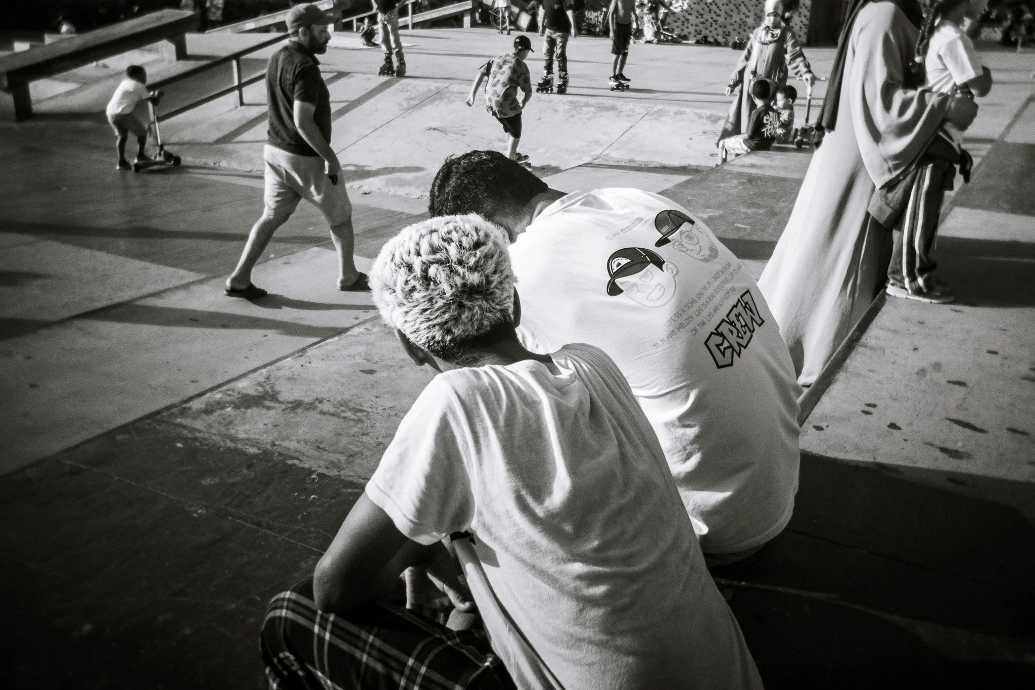 Black-and-white photo of people skating and socializing at an outdoor skate park, with two men seated in the foreground.