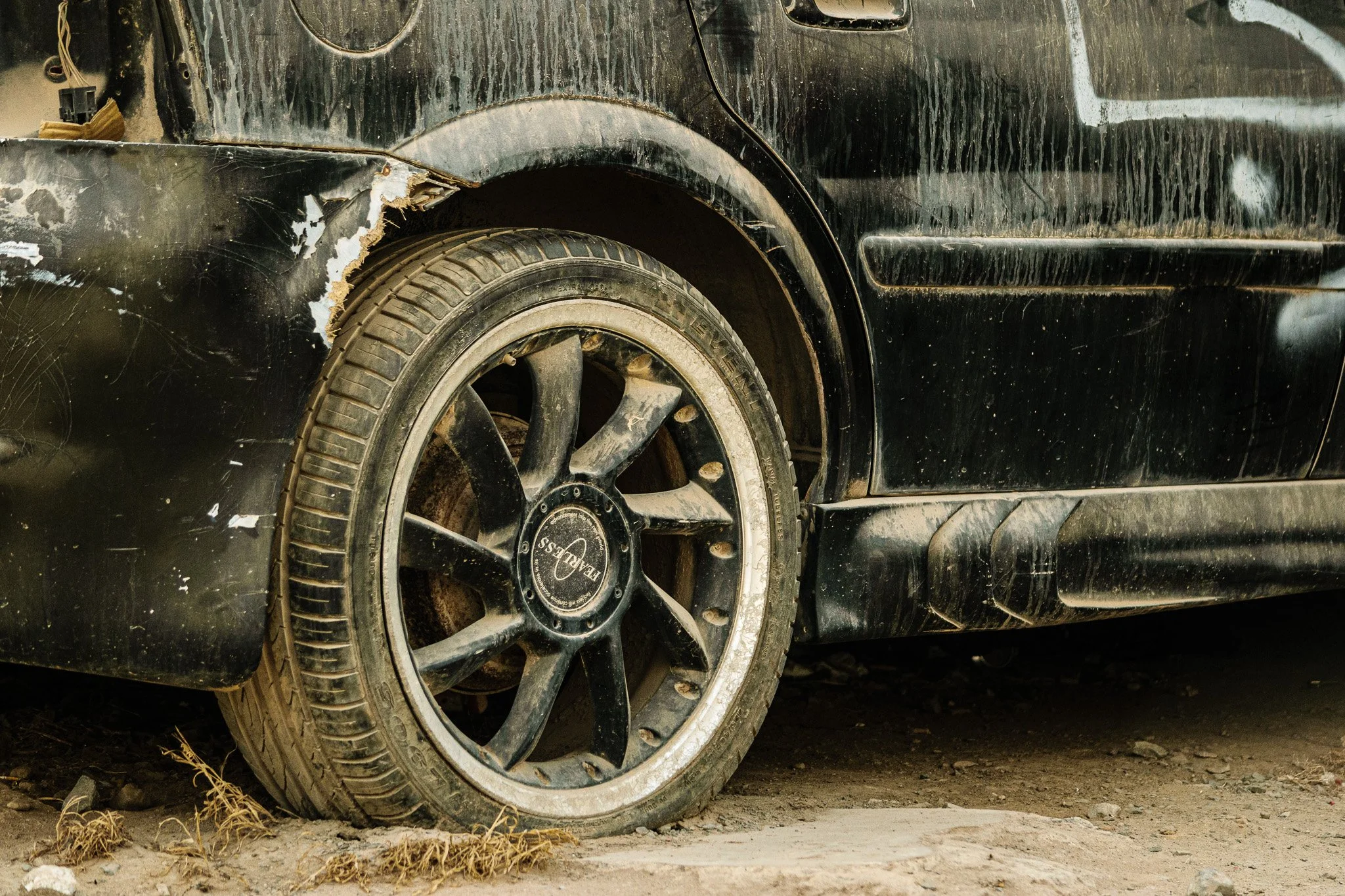 Close-up of a black car with one front tire showing dirt and mud, surrounded by scratches, dirt marks, and a damaged area near the wheel well.
