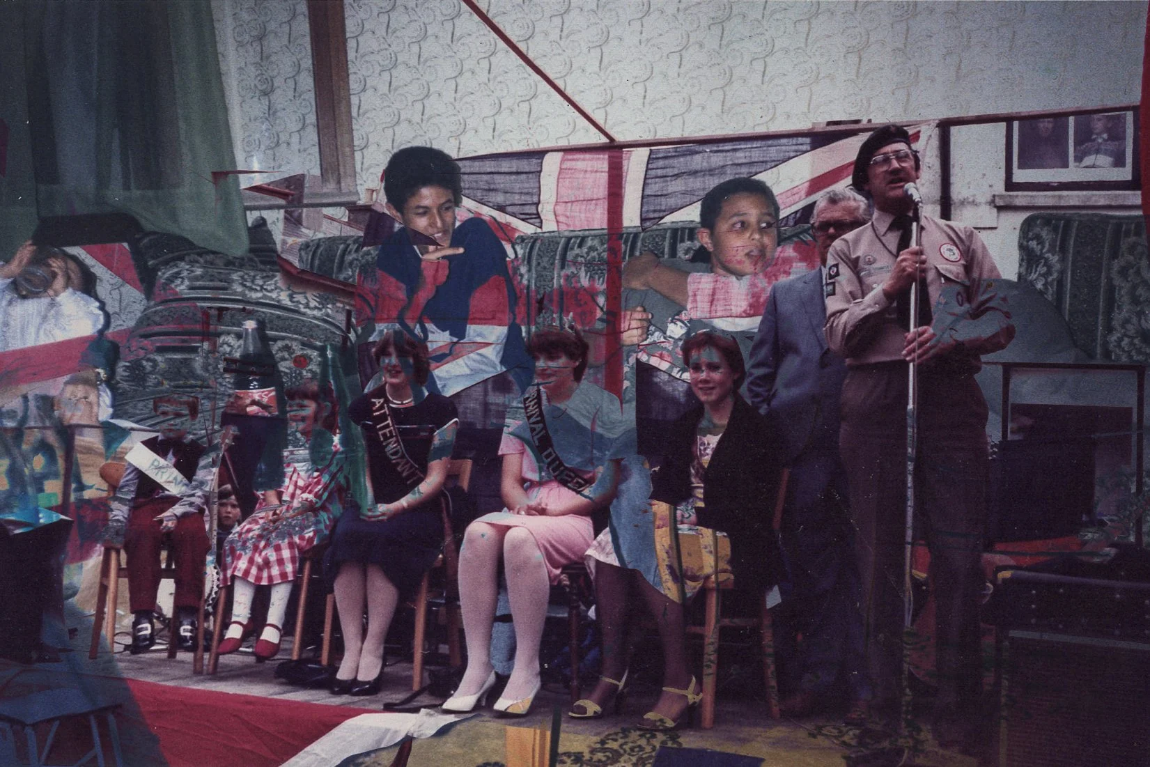 A group of children in costumes and sashes, sitting and standing on a stage during a performance or pageant, with an adult speaking into a microphone, in a room decorated with photos and fabric drapes.
