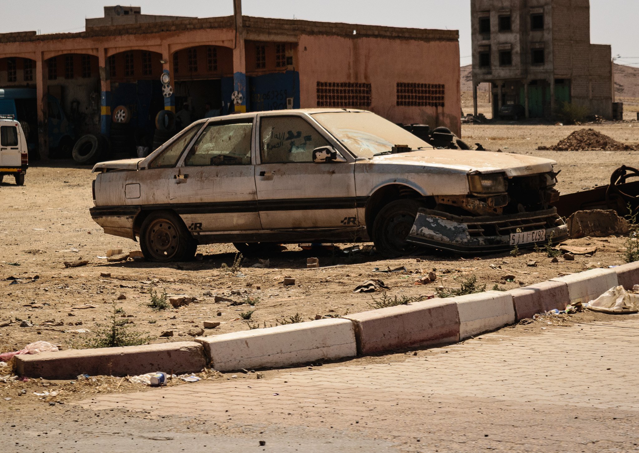 A dusty, abandoned car in a barren, desert-like area with scattered trash and debris. In the background, there are dilapidated buildings, one with a pinkish facade and another unfinished concrete structure. The sky appears clear but hazy.