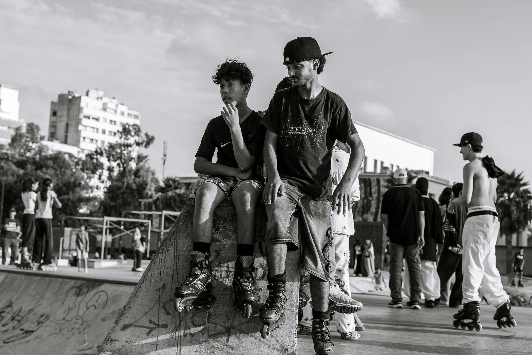 Black-and-white photo of young inline skaters gathered at an outdoor skate park, with two seated on a graffiti-covered ramp and others skating in the background.