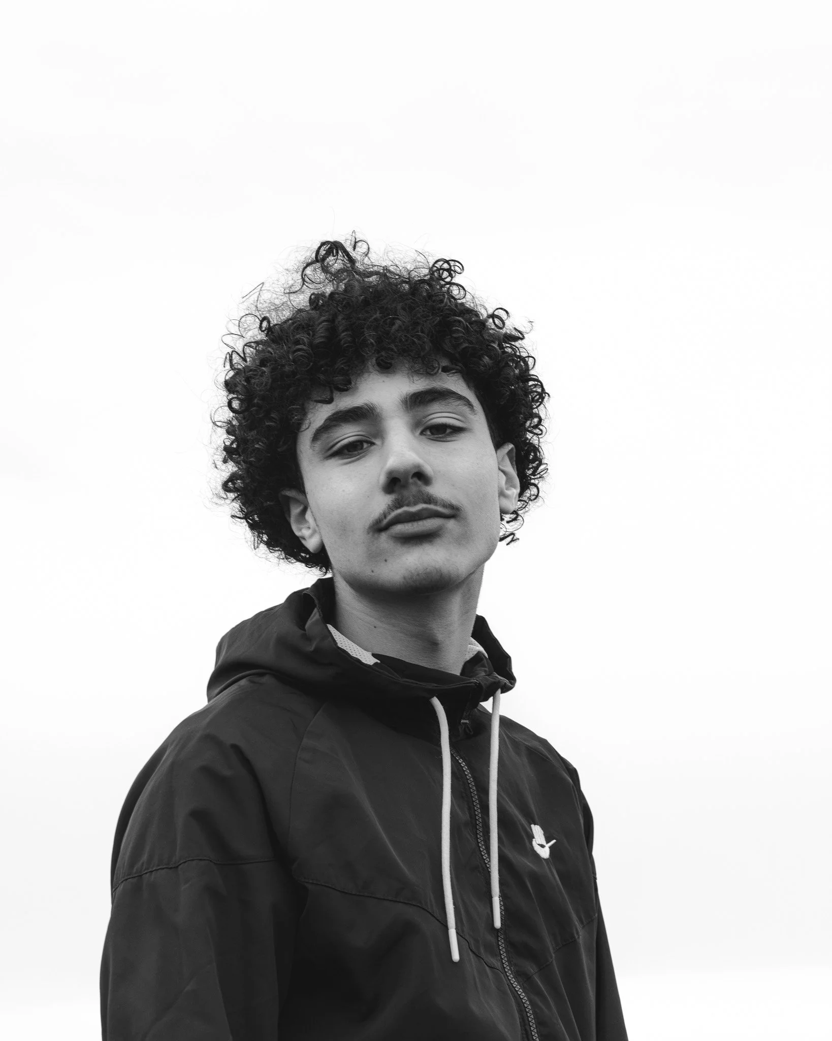 Black and white photograph of a young man with curly hair wearing a jacket with a zipper and a small logo. The background is plain and white.
