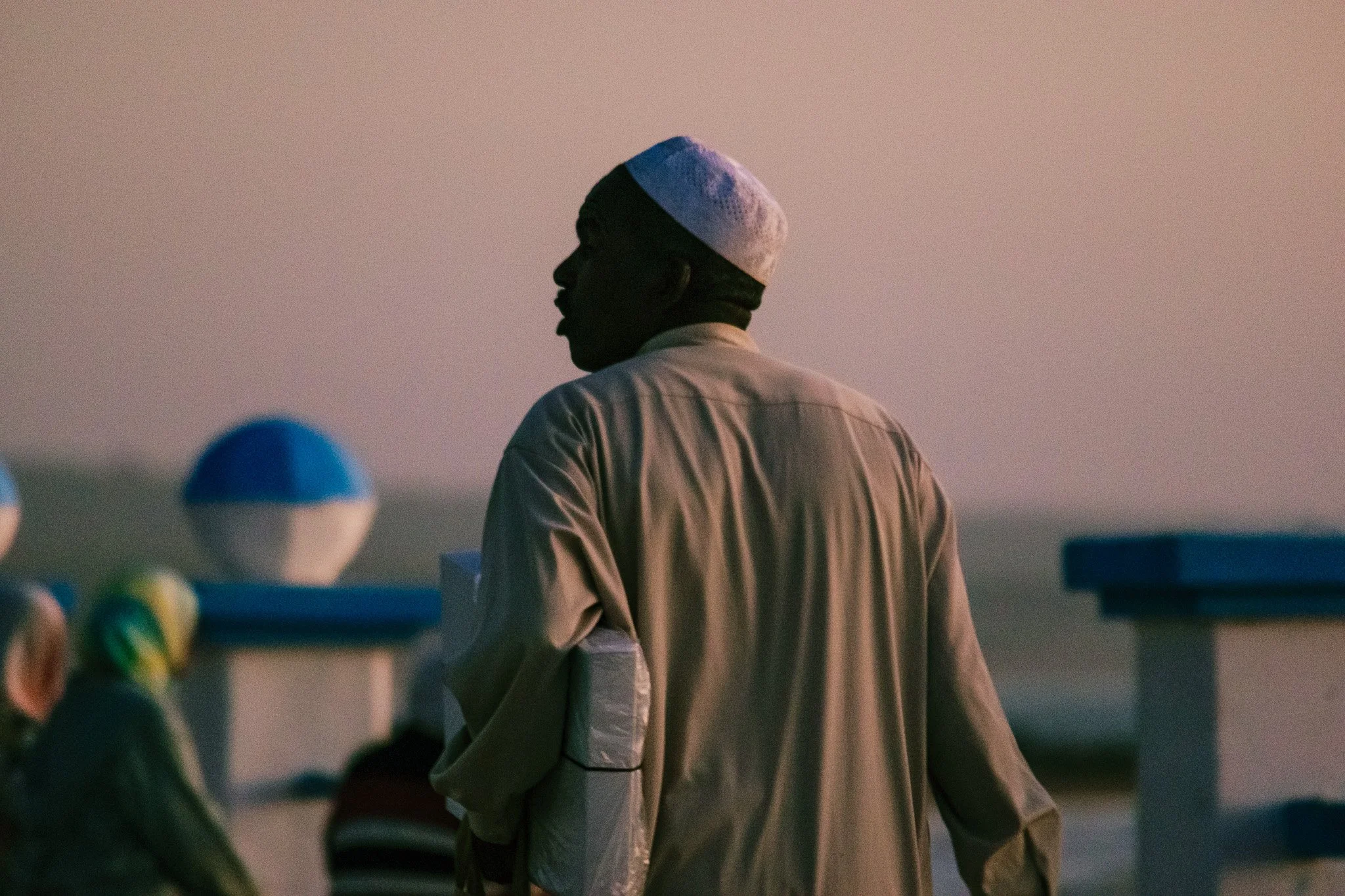 A man in traditional Islamic attire, including a white cap and a beige robe, standing outdoors during sunset.