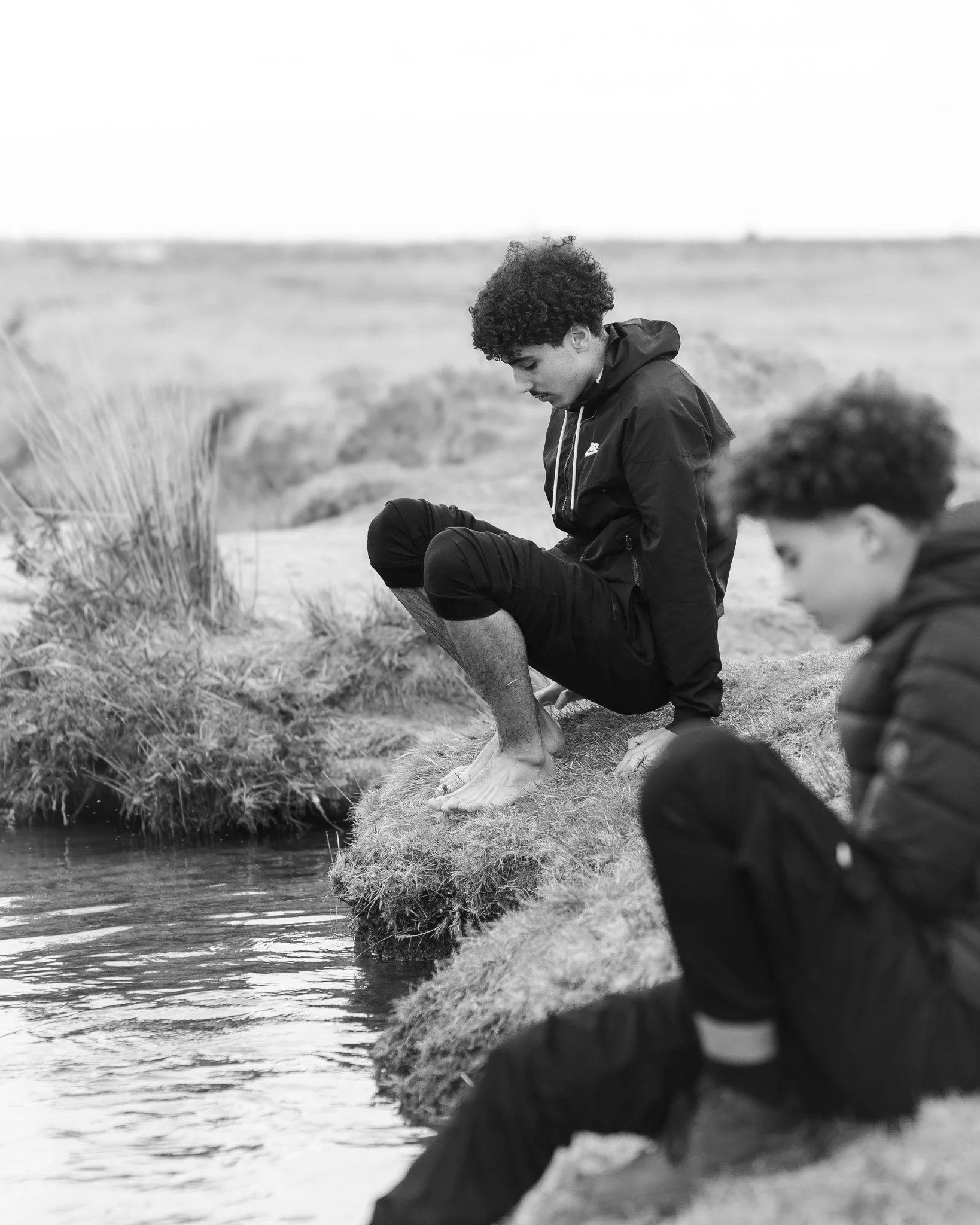 A black and white photo of a young man with curly hair sitting on a grassy bank by a body of water, looking down with a contemplative expression. There are other people partially visible in the foreground, out of focus.