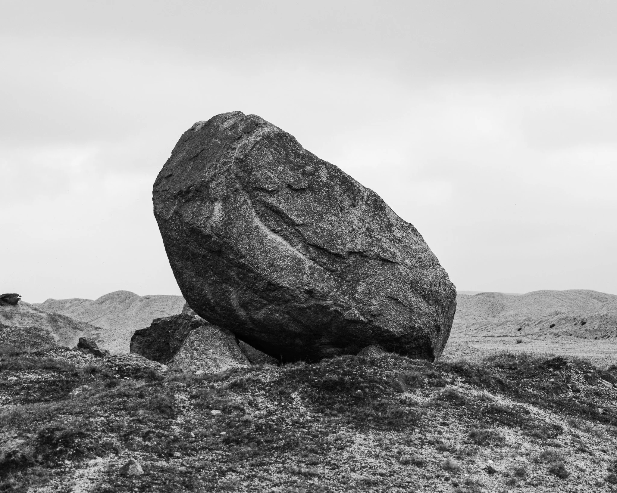 A large balancing rock formation in a desert landscape, with rugged terrain and overcast sky.