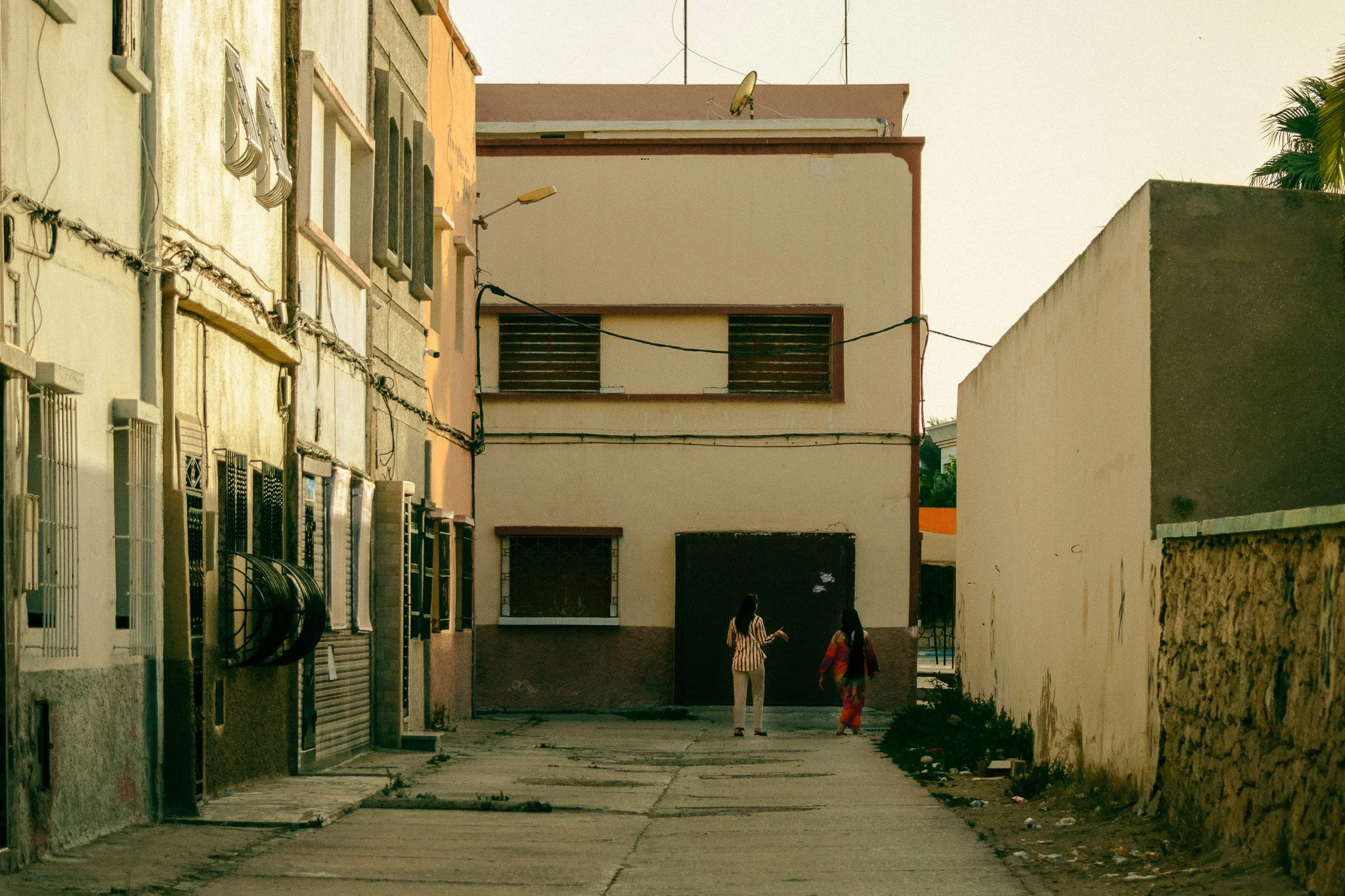 A narrow alleyway between residential buildings, with two women walking and talking, one in a striped top and the other in colorful traditional attire. The buildings have barred windows and various pipes.