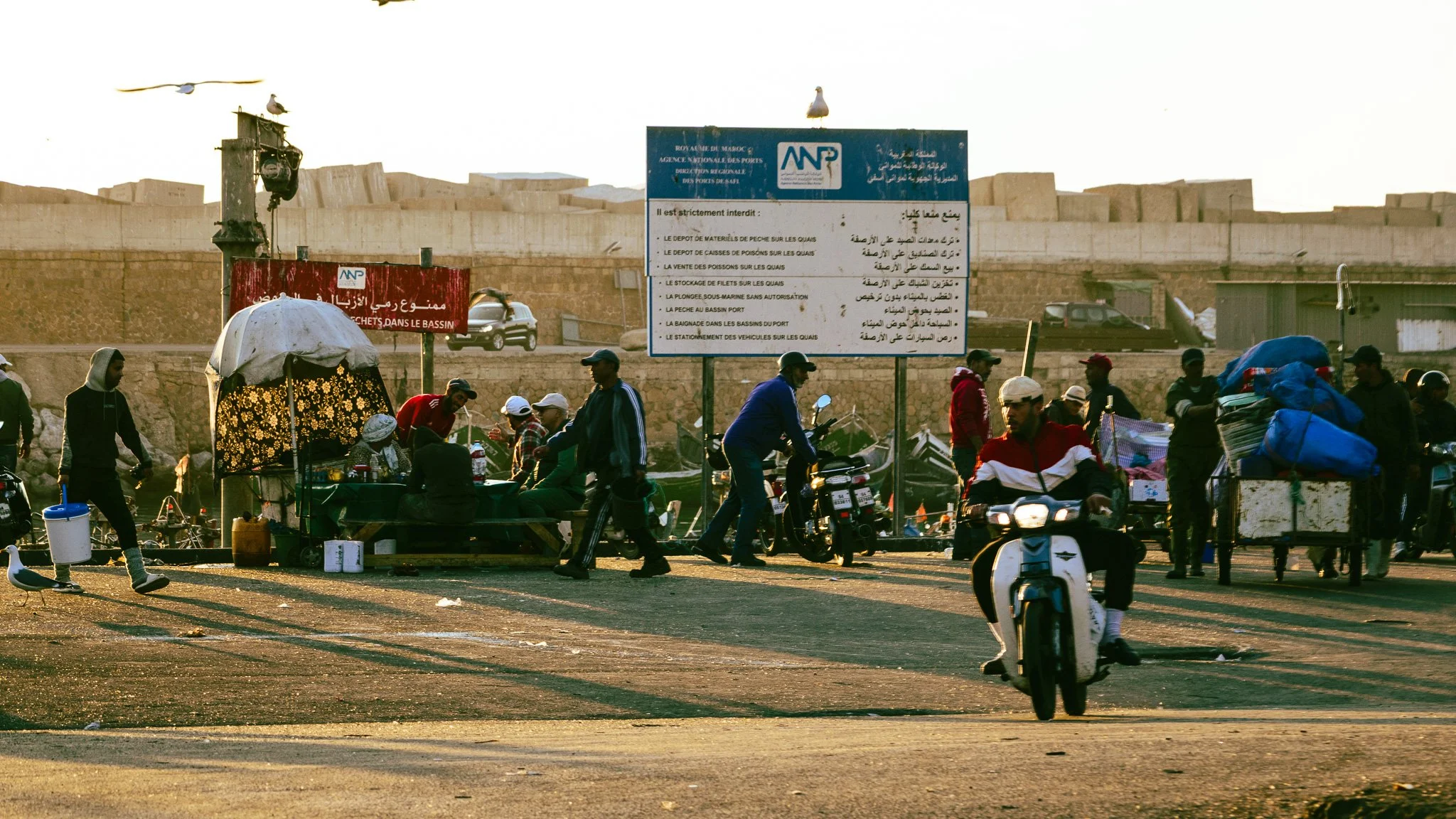 Street scene with people walking and riding scooters, vendor booth with umbrella, large signs in Arabic and French, and a large wall or structure in the background.