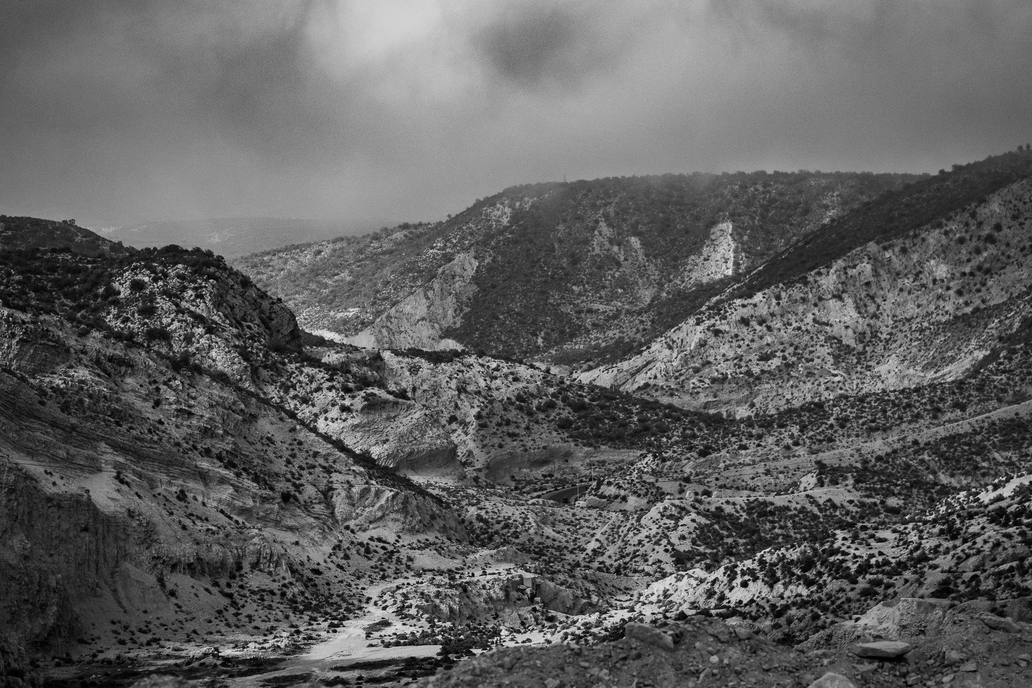 A black and white photograph of a mountainous landscape with steep, rugged terrain and scattered vegetation.
