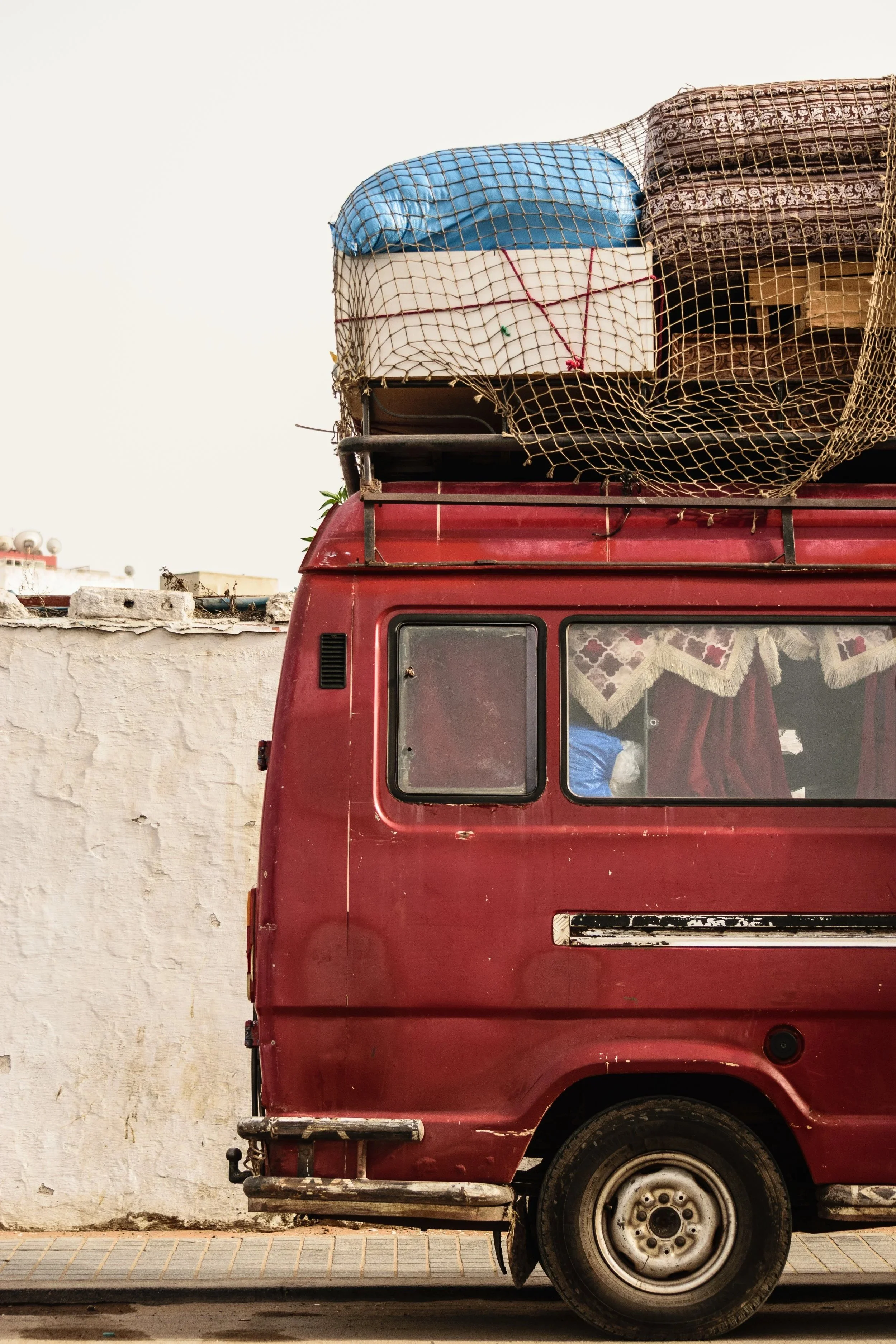A red van parked next to a white wall with items loaded on the roof, including a blue mattress, a box, and blankets, all secured with netting.
