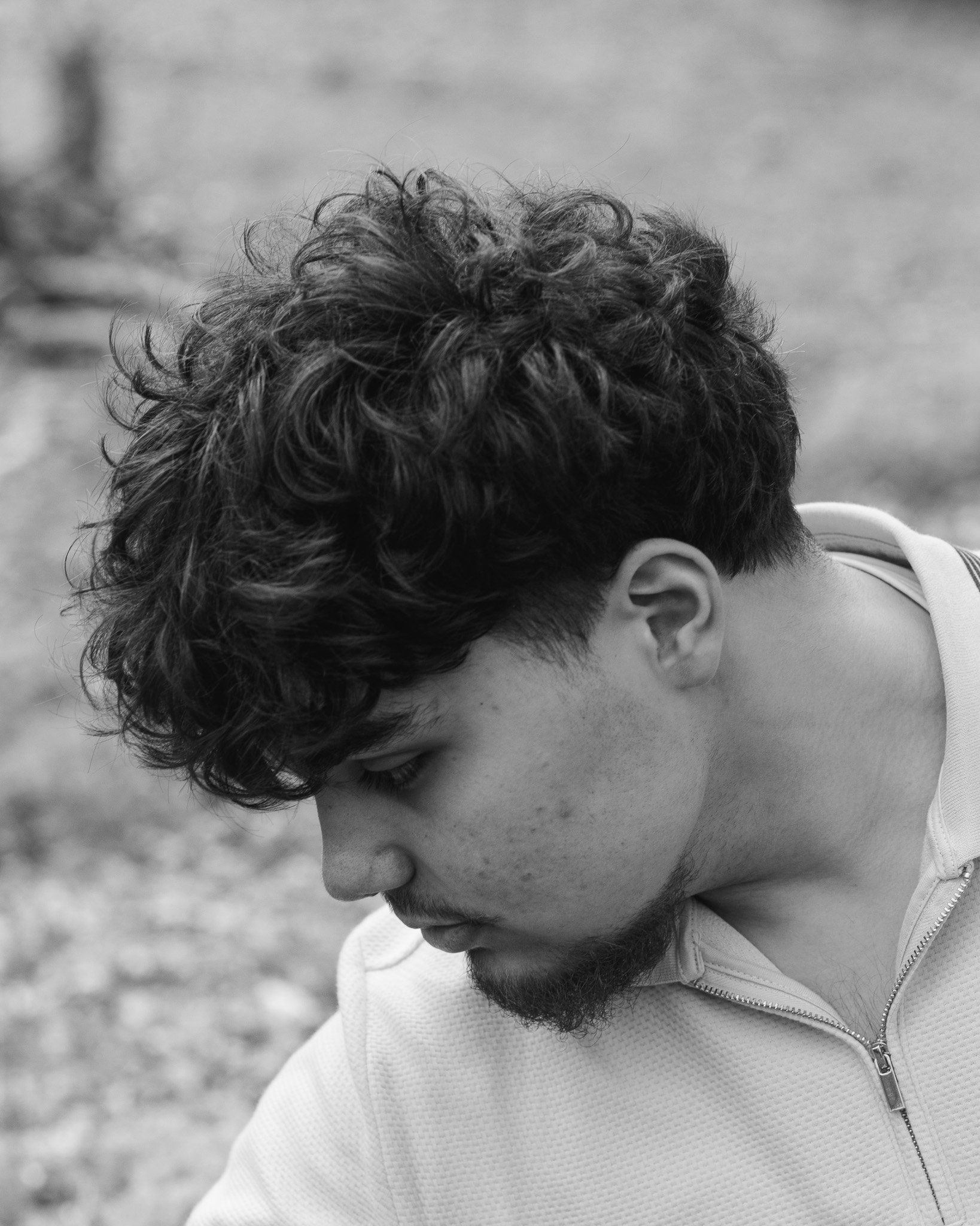 Close-up of a young man with curly hair and a beard, looking downward, wearing a zip-up jacket.