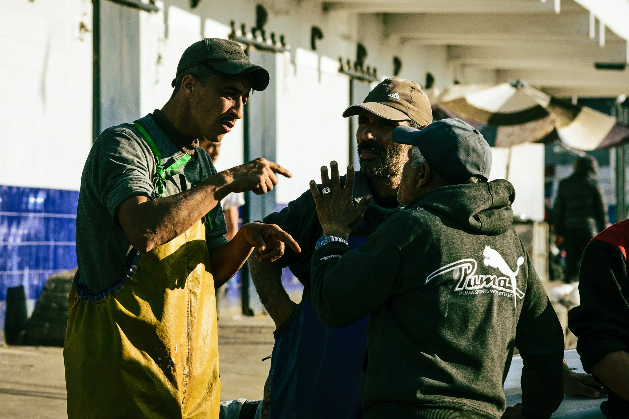 Three men engaged in conversation outdoors, two of them wearing caps and casual clothing, with a background of white buildings and umbrellas.