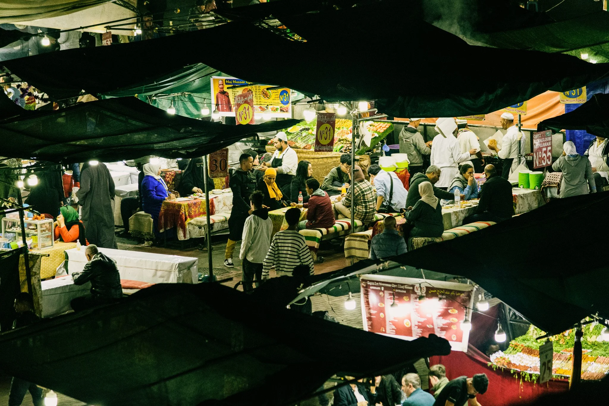 Night market scene with people shopping and eating under tents, illuminated advertised food stalls, and a crowded atmosphere.