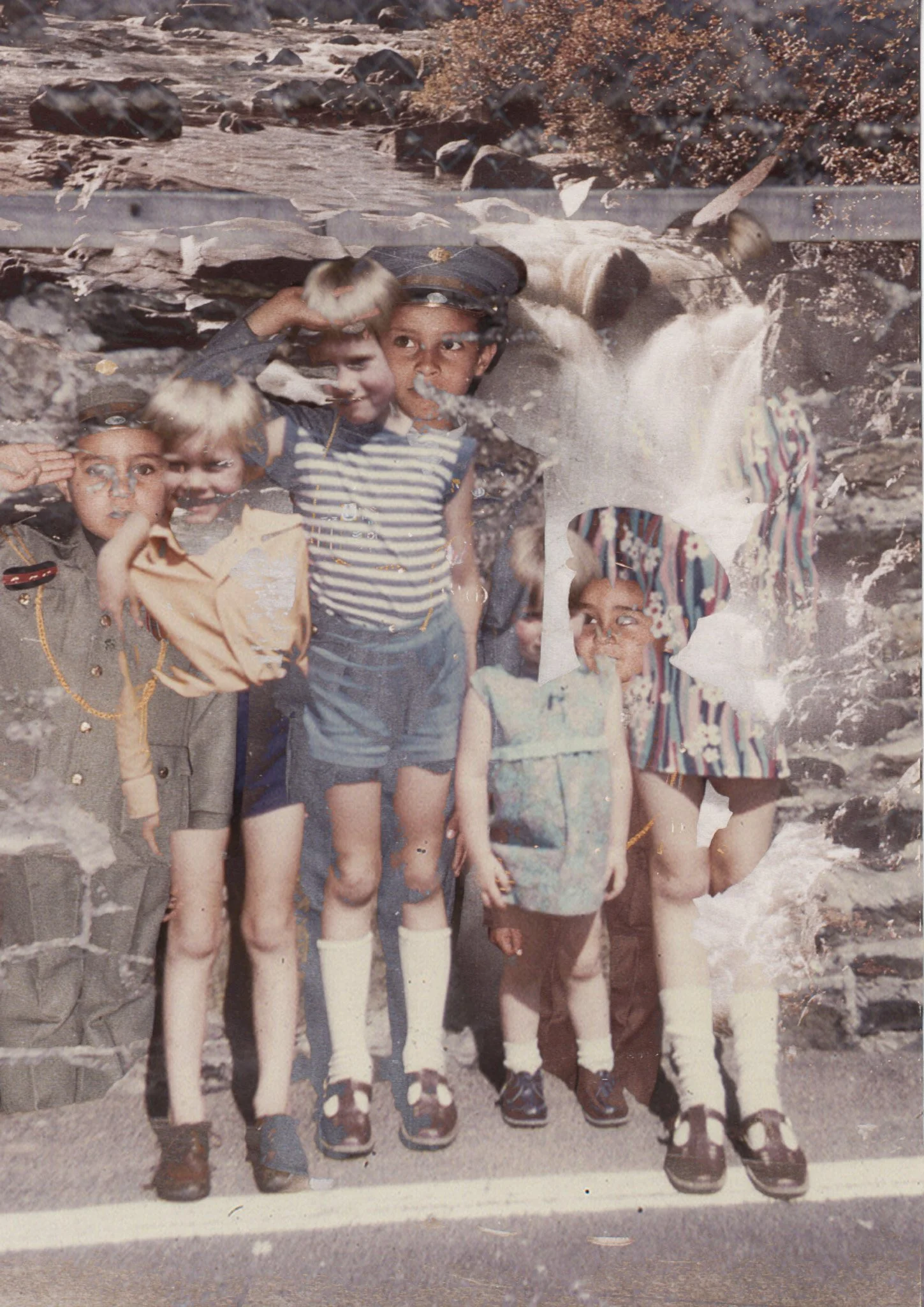 Group of children in scout uniforms and casual clothing standing outdoors on a road, with rocky terrain and trees in the background.