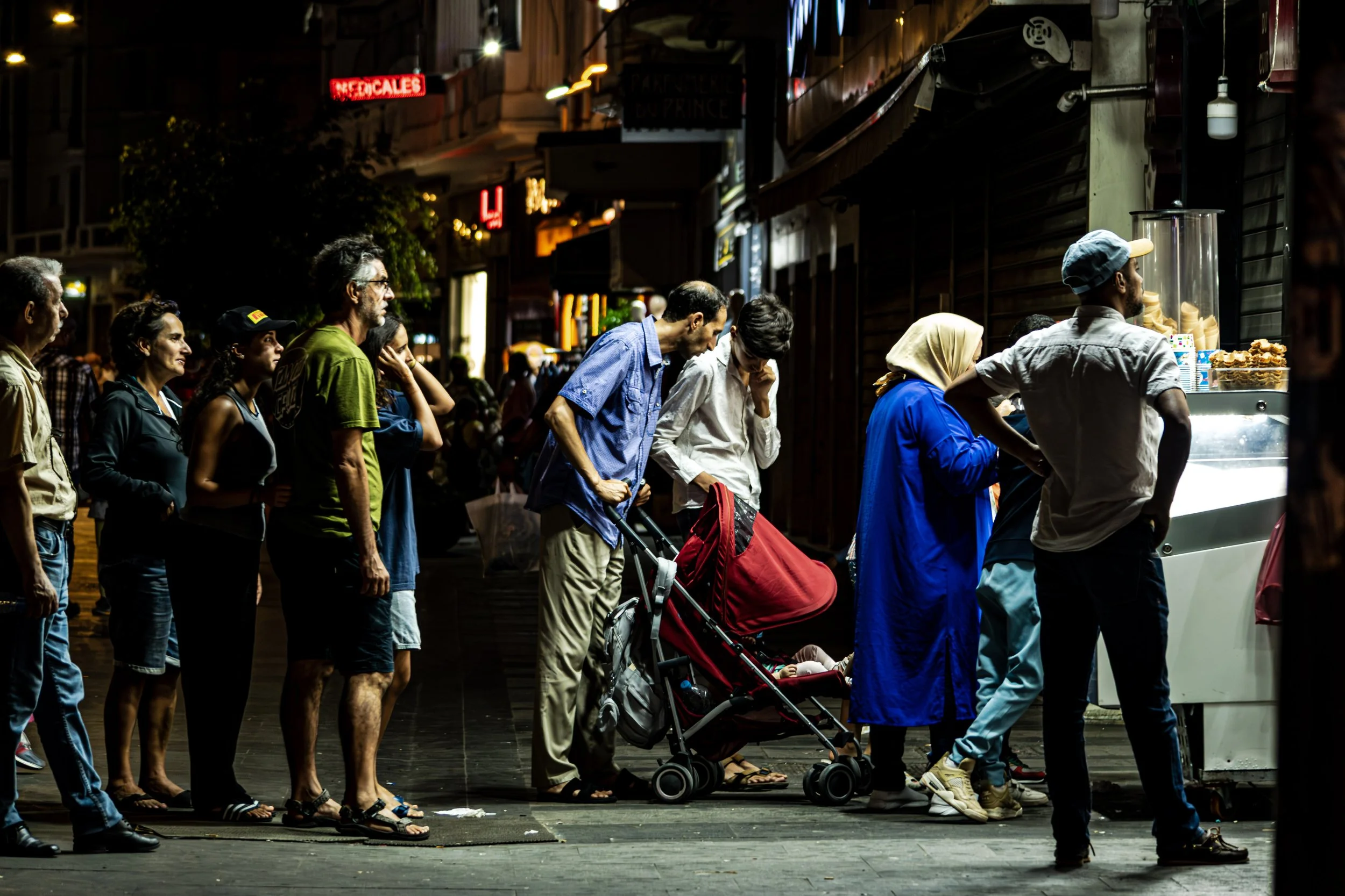 Line of diverse people standing outside at night, waiting for food at a food cart with a glass display of snacks amidst city signs and buildings.