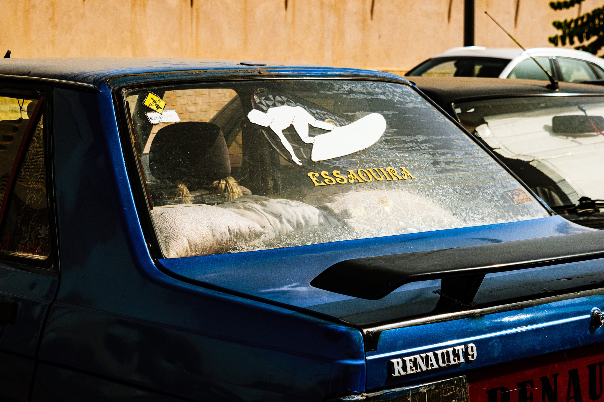 A close-up of a blue race car with a white emblem of a dog and the word 'ESSAOURIA' on the windshield. Two other cars are visible in the background.