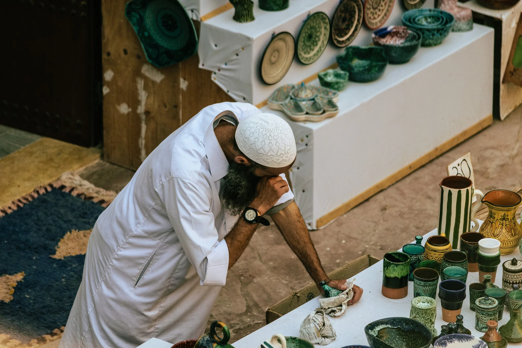 A man wearing a white shirt and a white cap looking down at pottery items on a table at an outdoor market.