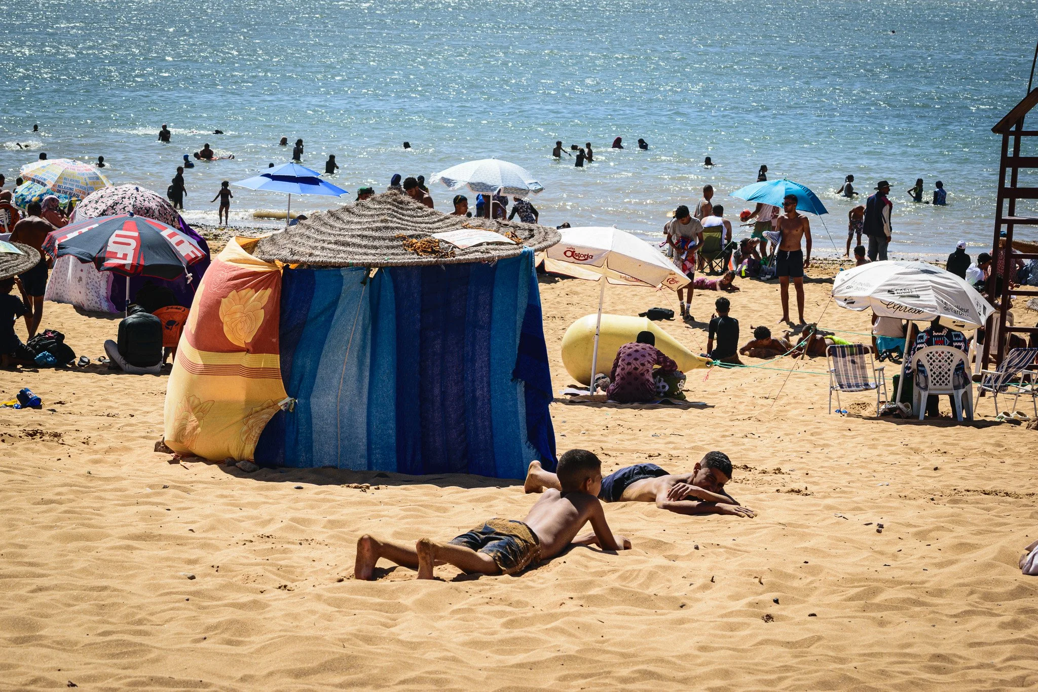 Crowded beach with umbrellas, straw huts, and people swimming, sunbathing, and relaxing on the sand.