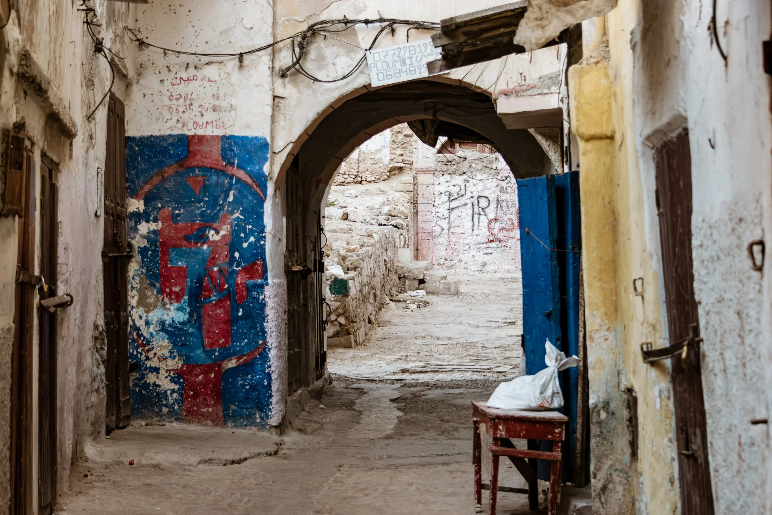 A weathered alleyway with old, chipped walls and graffiti. An arched doorway leads into a narrow street, with a small table and a plastic bag visible in the scene.