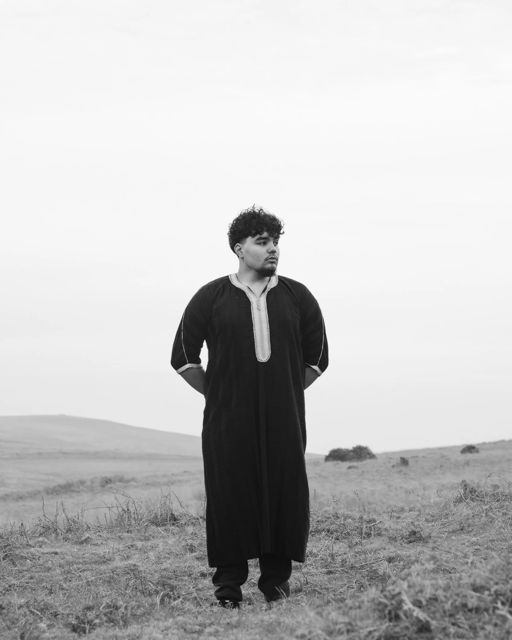 A man in traditional South Asian attire stands outdoors in a grassy field with an overcast sky, looking to the side.