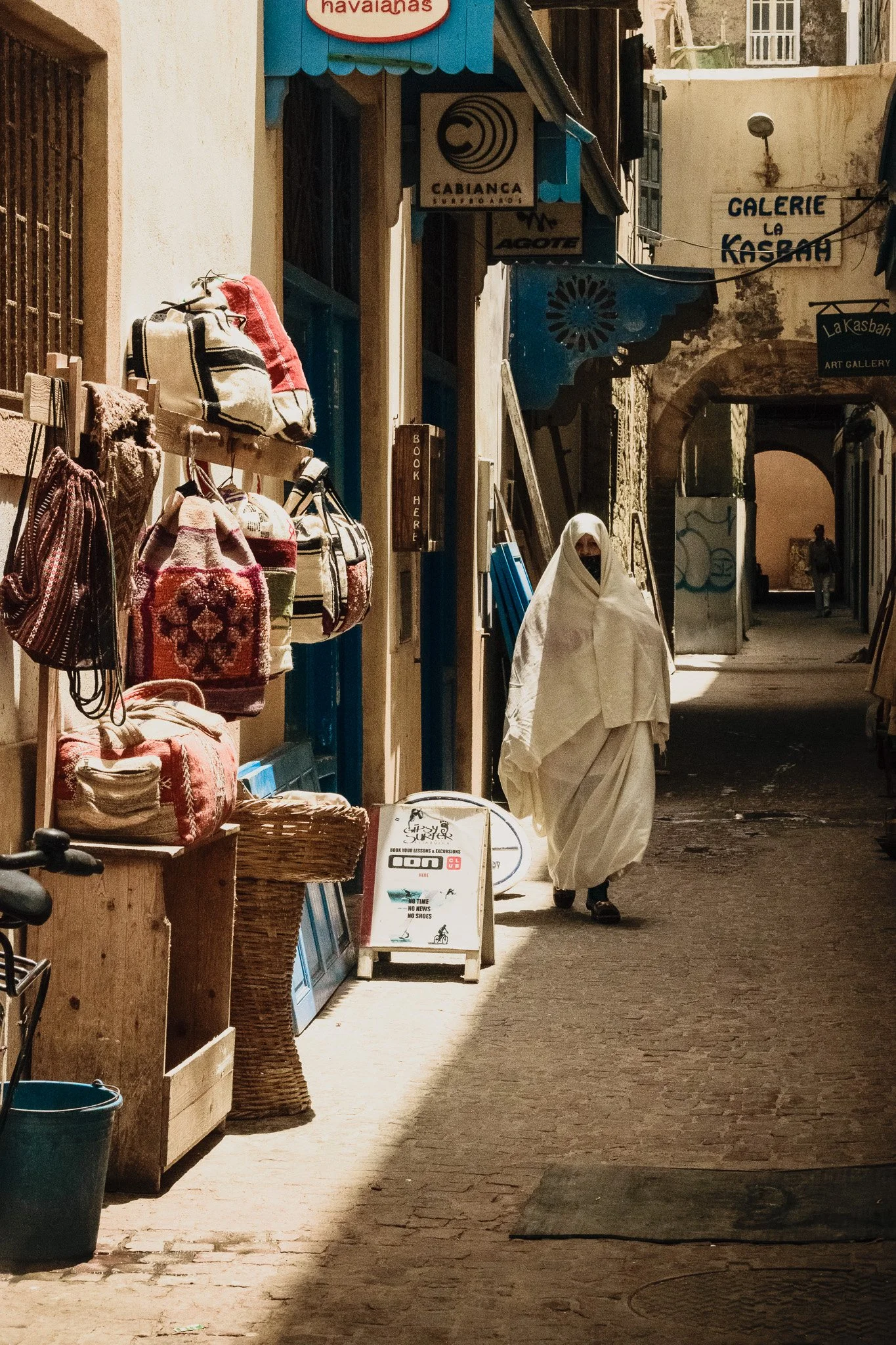 A narrow street with shops displaying bags on the left, people walking and browsing, and signs indicating art galleries and a bookstore. One person is wrapped in white cloth walking down the street.