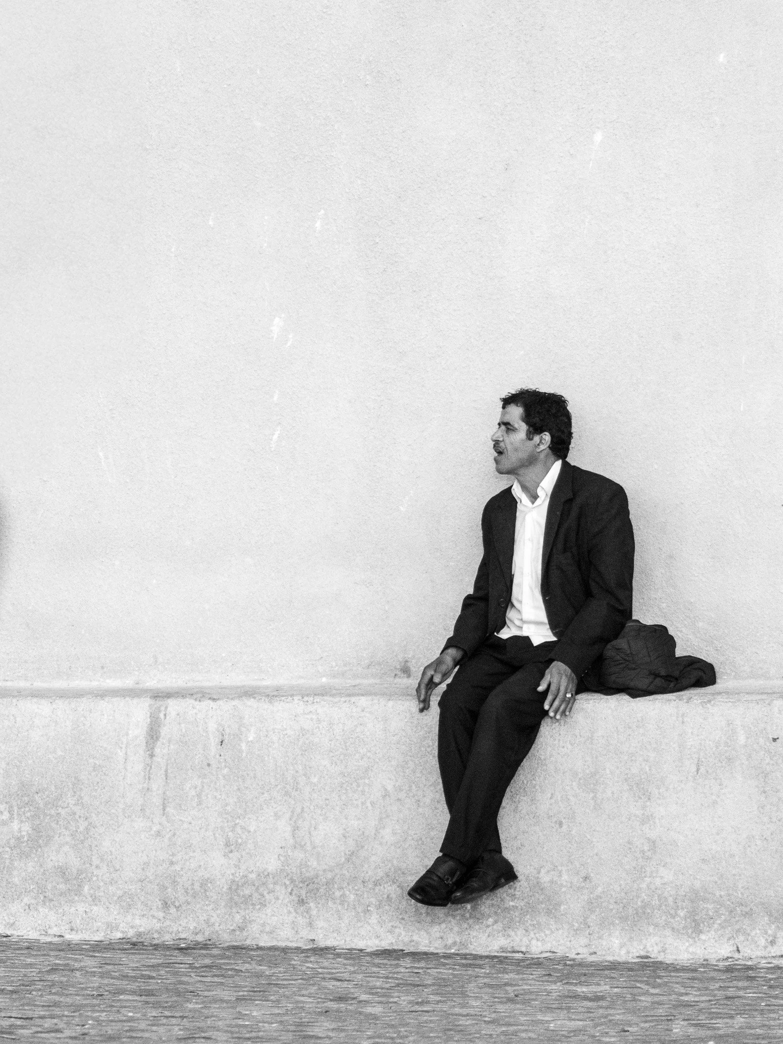 A man in a suit sitting on a concrete ledge with a backpack beside him, facing to the right, against a plain white wall.