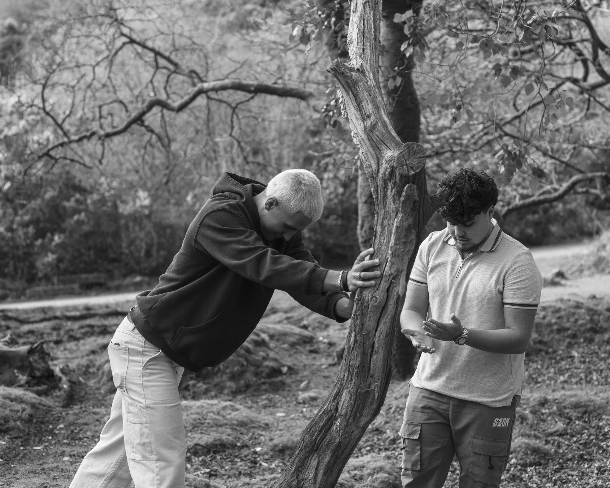 Two young men are outdoors, leaning over a large, gnarled tree trunk, looking at an object in one of the man's hands. The scene is in black and white and has a background of tree branches and natural surroundings.