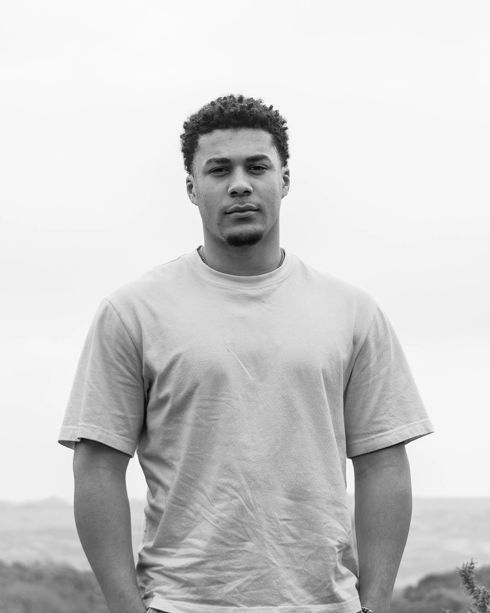 Black and white photo of a young man with curly hair and a goatee, standing outdoors with a blurred landscape background, wearing a plain T-shirt.