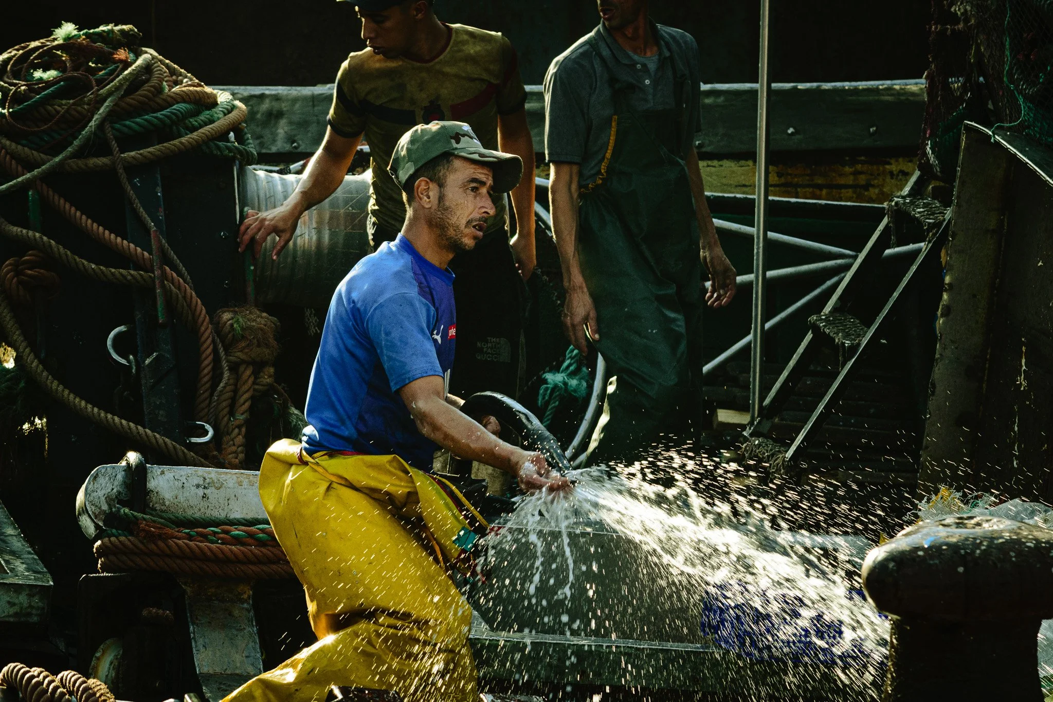 A man in yellow waterproof gear and a cap is sitting on a boat, handling a rope, while two other men stand nearby, with one touching thick ropes and the other observing. They are on a boat filled with nautical equipment, and water is splashing as the