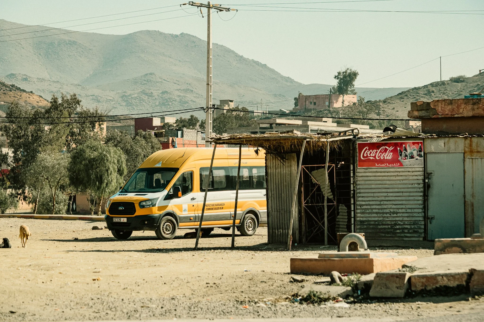 A yellow and white vehicle parked in a rural area with dry ground, a dog walking nearby, and simple structures. Mountains are visible in the background, and there is a Coca-Cola sign on one of the structures.
