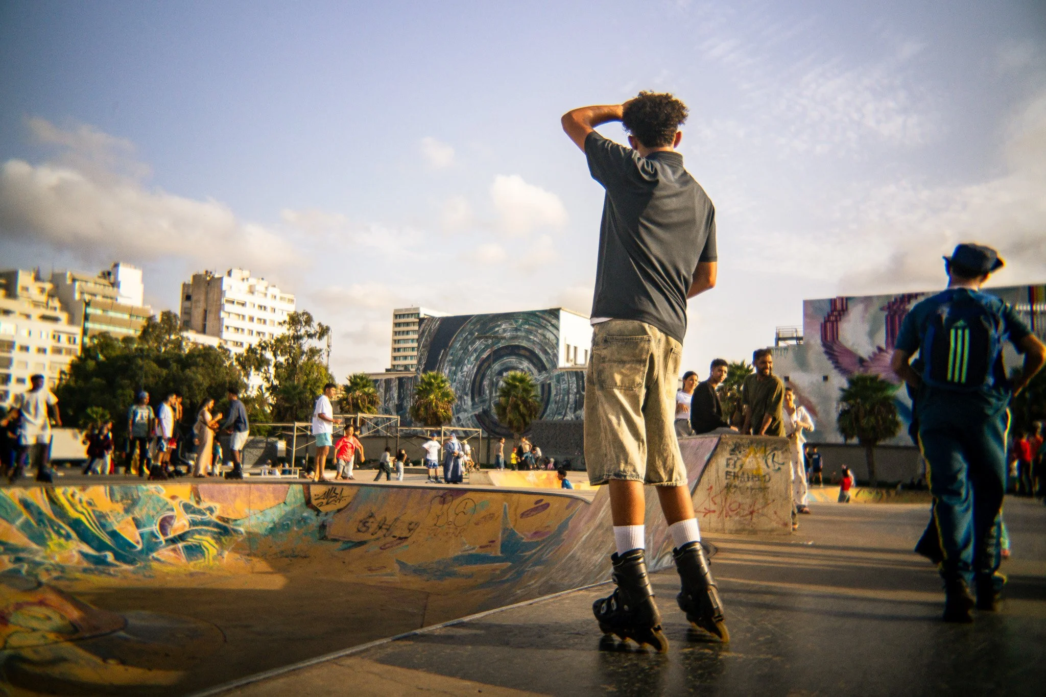 People rollerblading and skateboarding at a graffiti-covered urban skate park with city buildings in the background