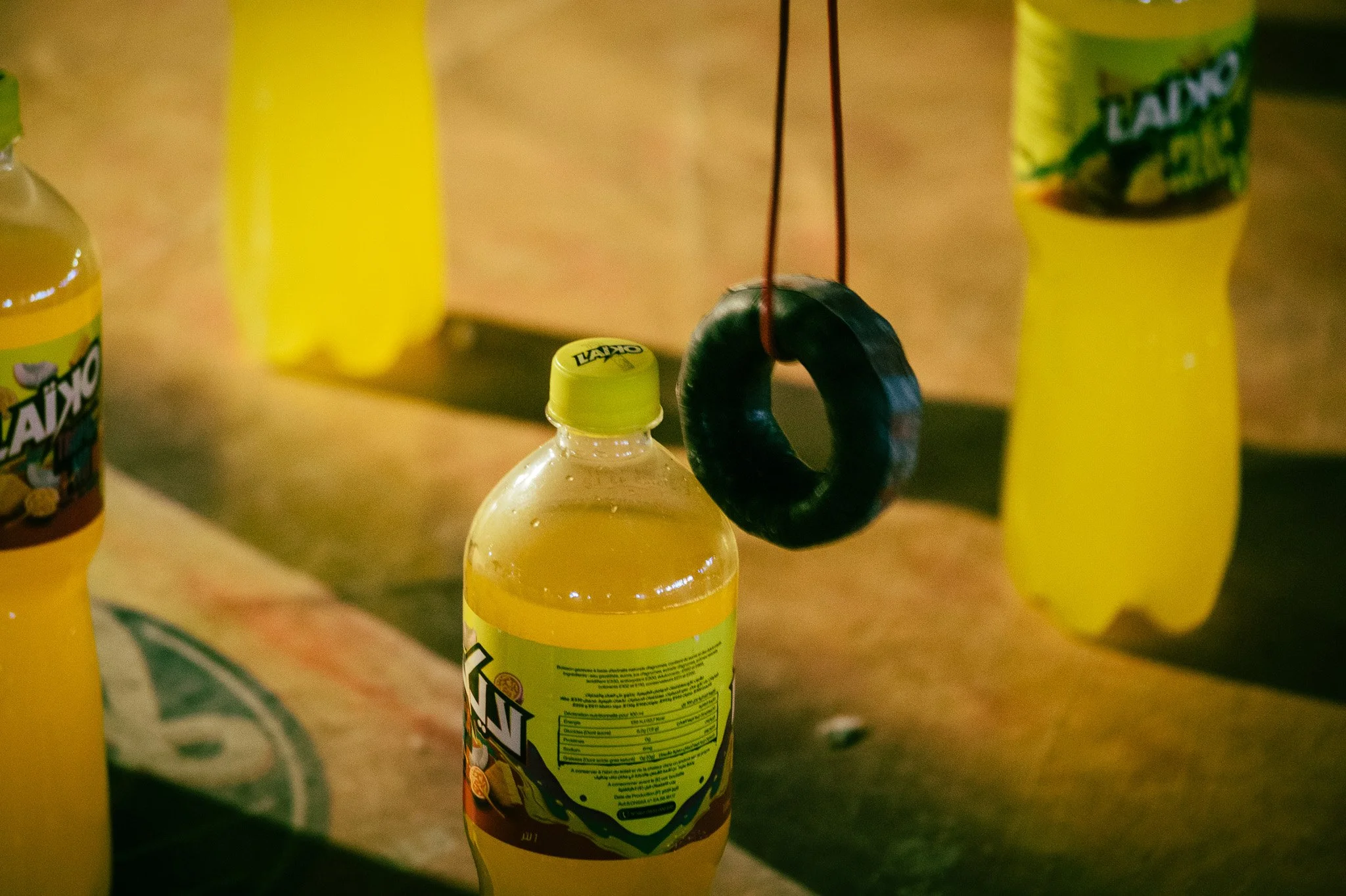 A yellow plastic bottle with a label, a black rubber ring hanging from red strings attached to the cap, and two other yellow bottles in the background.