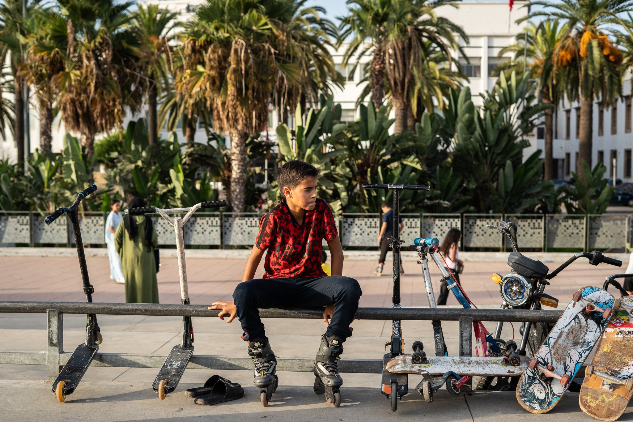 Boy wearing inline skates sits on a skatepark rail among scooters and skateboards, with palm trees and a building behind him.