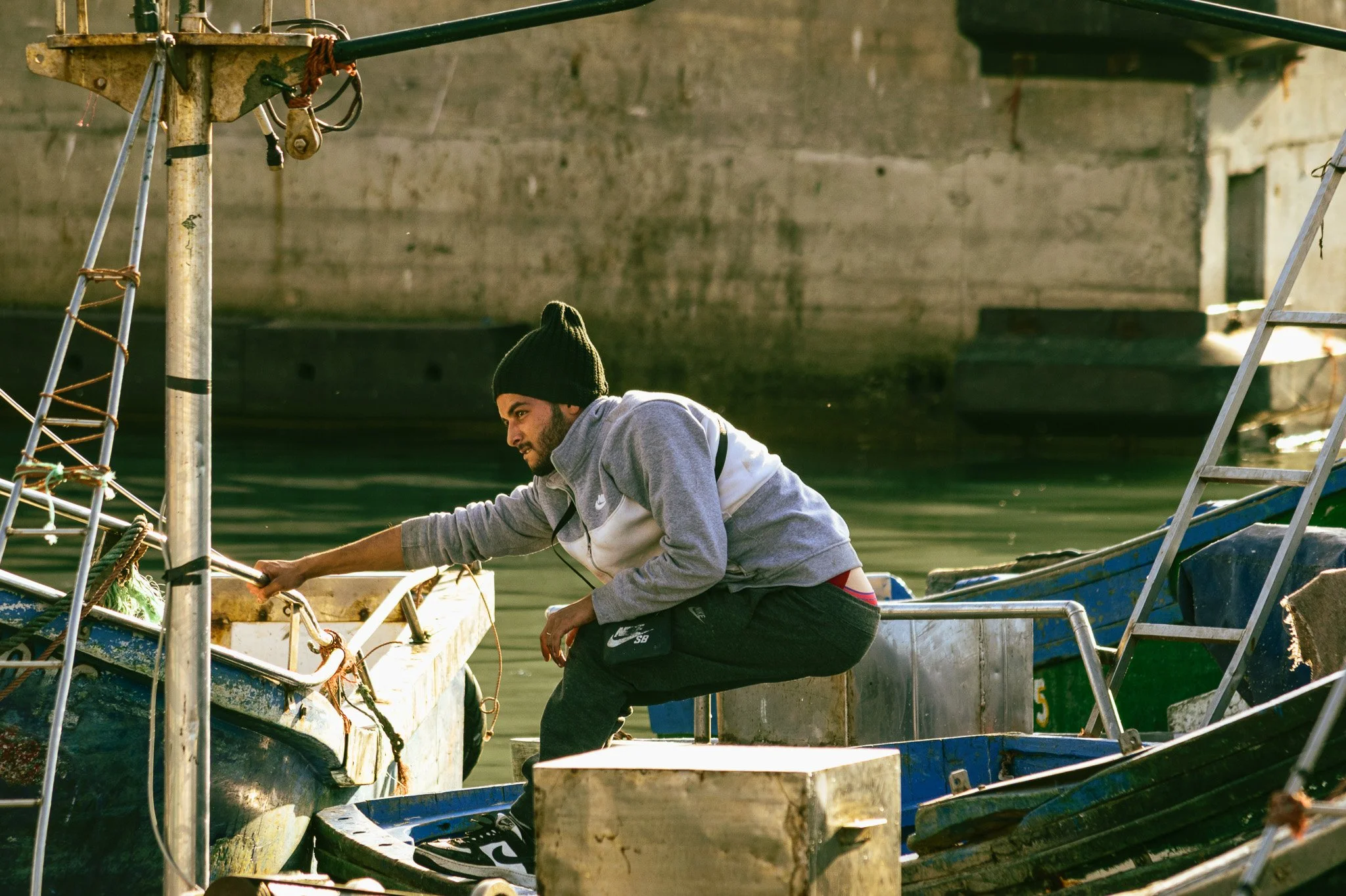 A man wearing a black beanie, gray hoodie, and black Nike shorts working on a boat near the water, with concrete structures in the background.