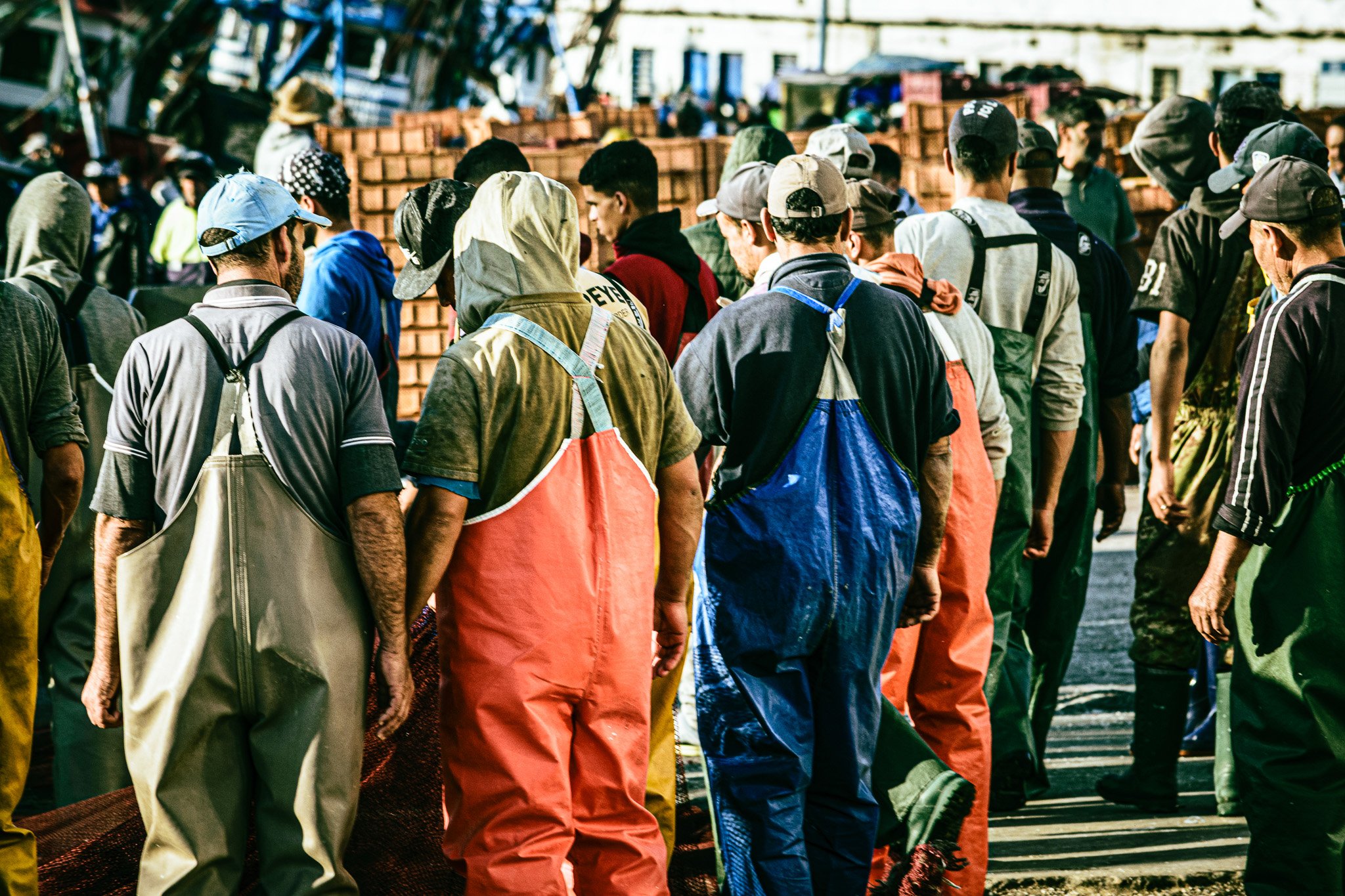 A group of people standing in line outdoors, some wearing caps and hoodies, with stacks of bricks in the background.