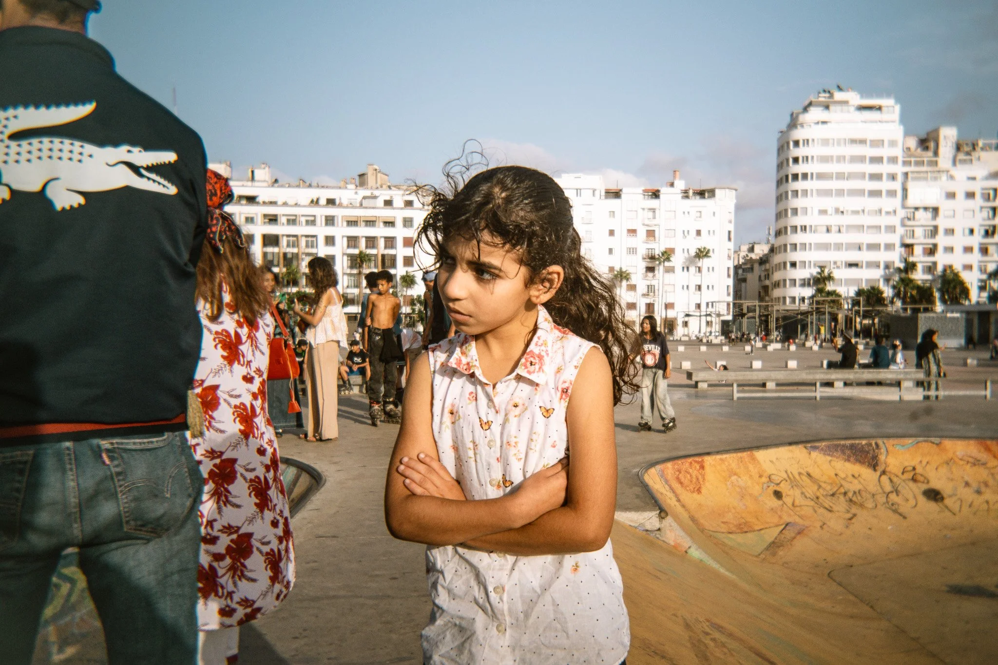 Young girl standing with folded arms beside a graffiti-covered skate bowl in an urban plaza, surrounded by people and city buildings.