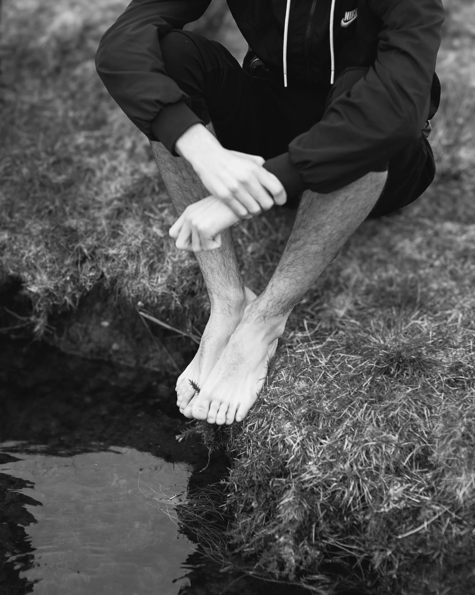 A person wearing a Nike jacket is kneeling beside a body of water, holding what appears to be a dead or injured bird.