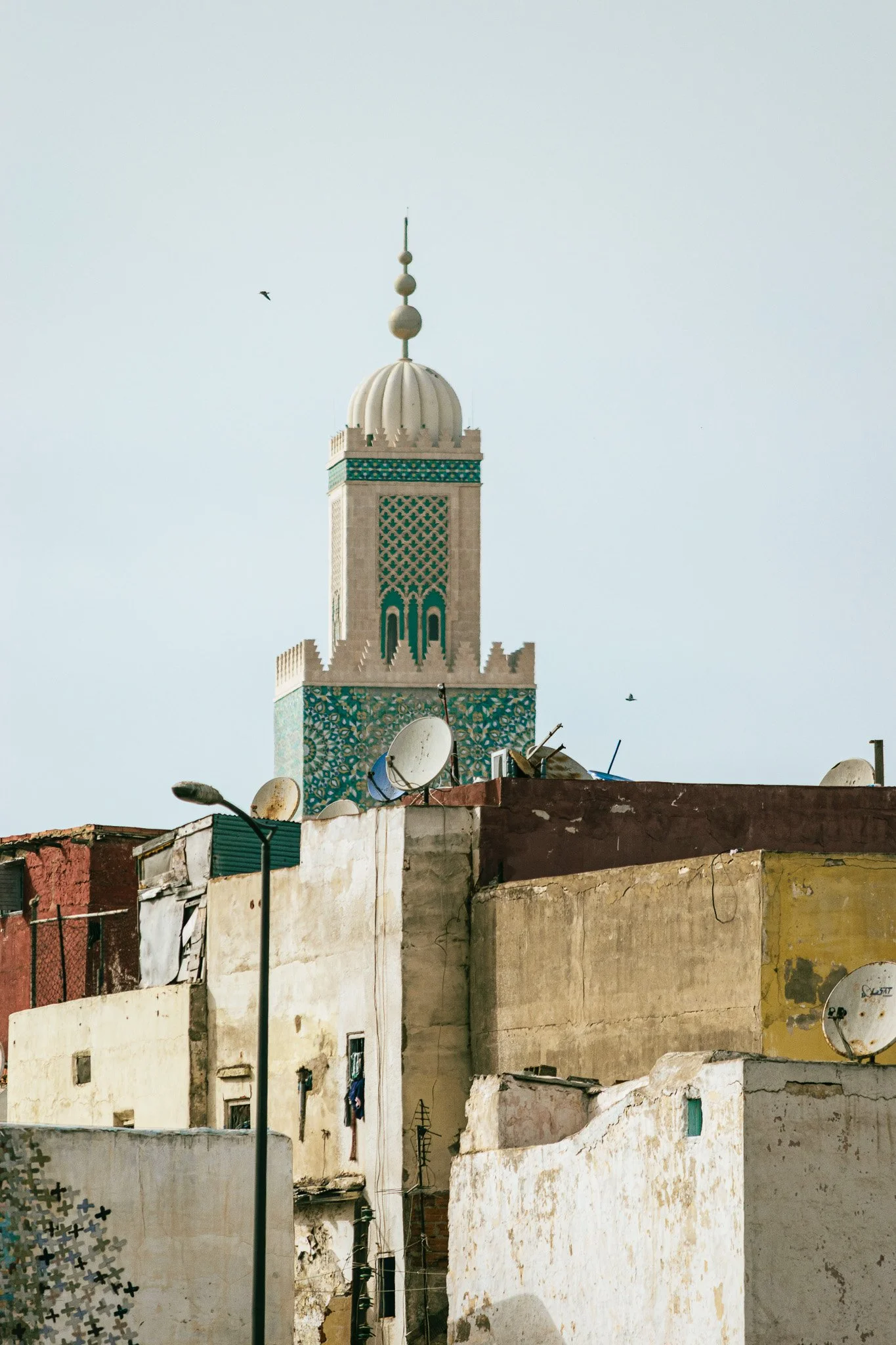 A cityscape with a tall minaret featuring a domed top and satellite dishes, surrounded by various old buildings and street lamps under a clear sky.