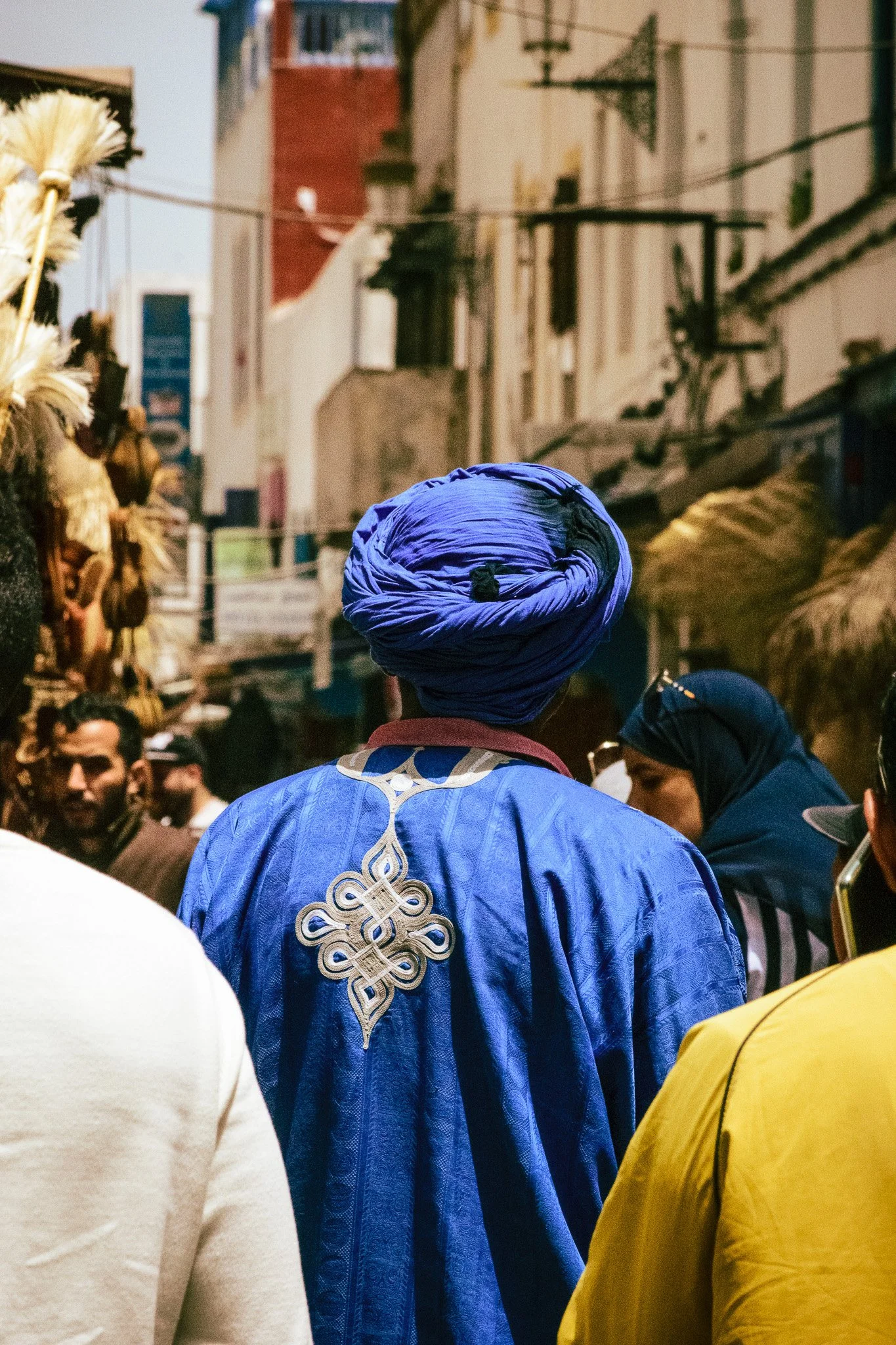 A person wearing a traditional blue outfit with intricate gold embroidery on the back and a matching blue turban, standing in a busy outdoor marketplace with several other people around.