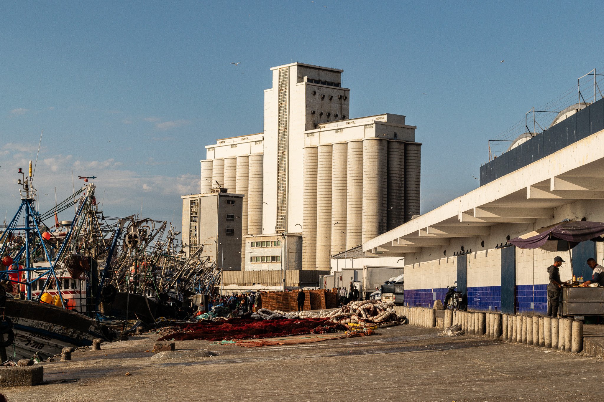 A harbor with fishing boats docked on the left and a large white industrial building with tall cylindrical silos in the background under a blue sky. There are people walking and working along the waterfront.