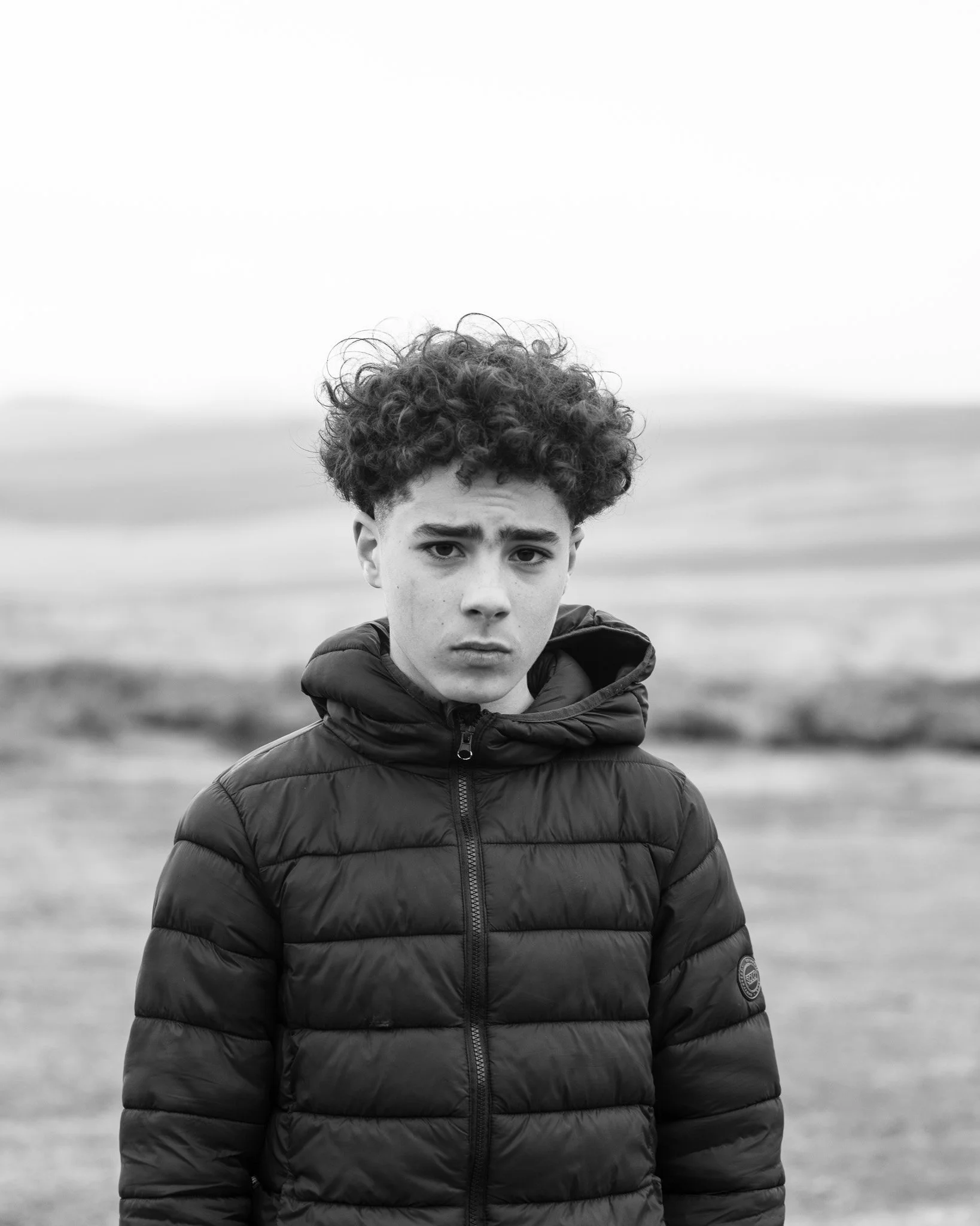 A young man with curly hair and a serious expression wearing a padded jacket outdoors, with a blurred landscape background in black and white.