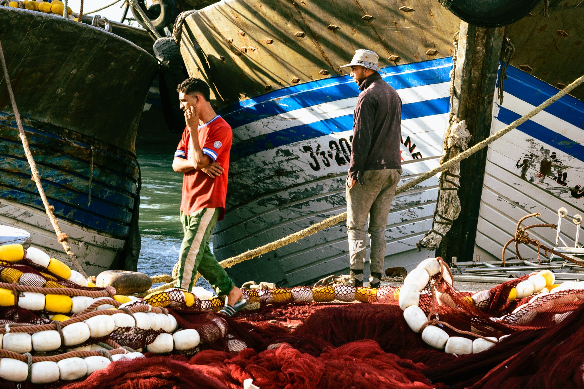 Two men walking on a boat docked at the harbor, with large boat hulls in the background and ropes, buoys, and fishing nets in the foreground.