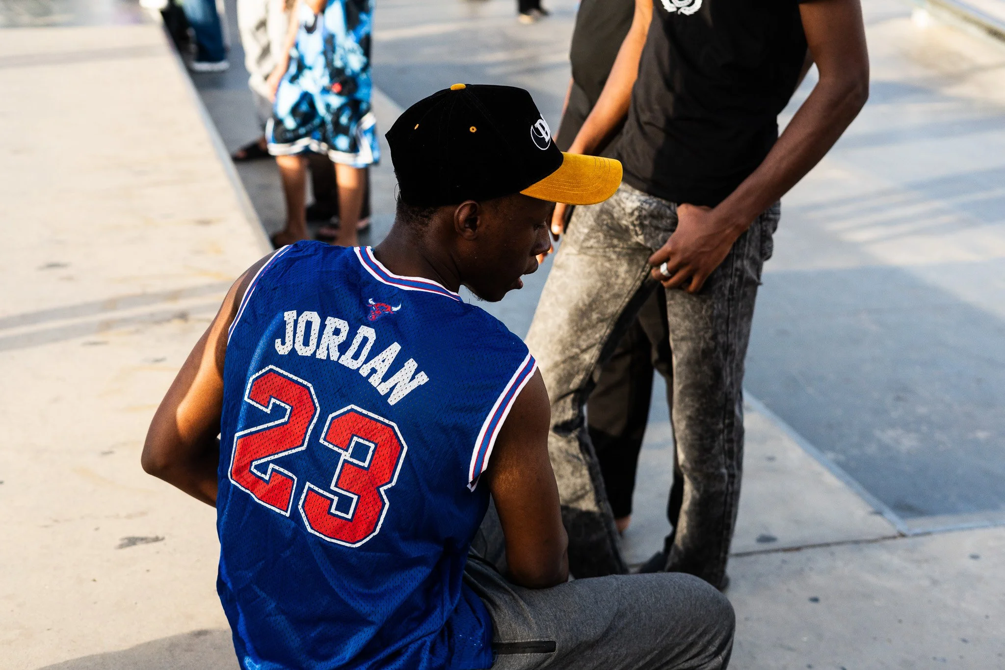 Man wearing a blue Jordan 23 basketball jersey and black-and-yellow cap sitting outdoors with people nearby