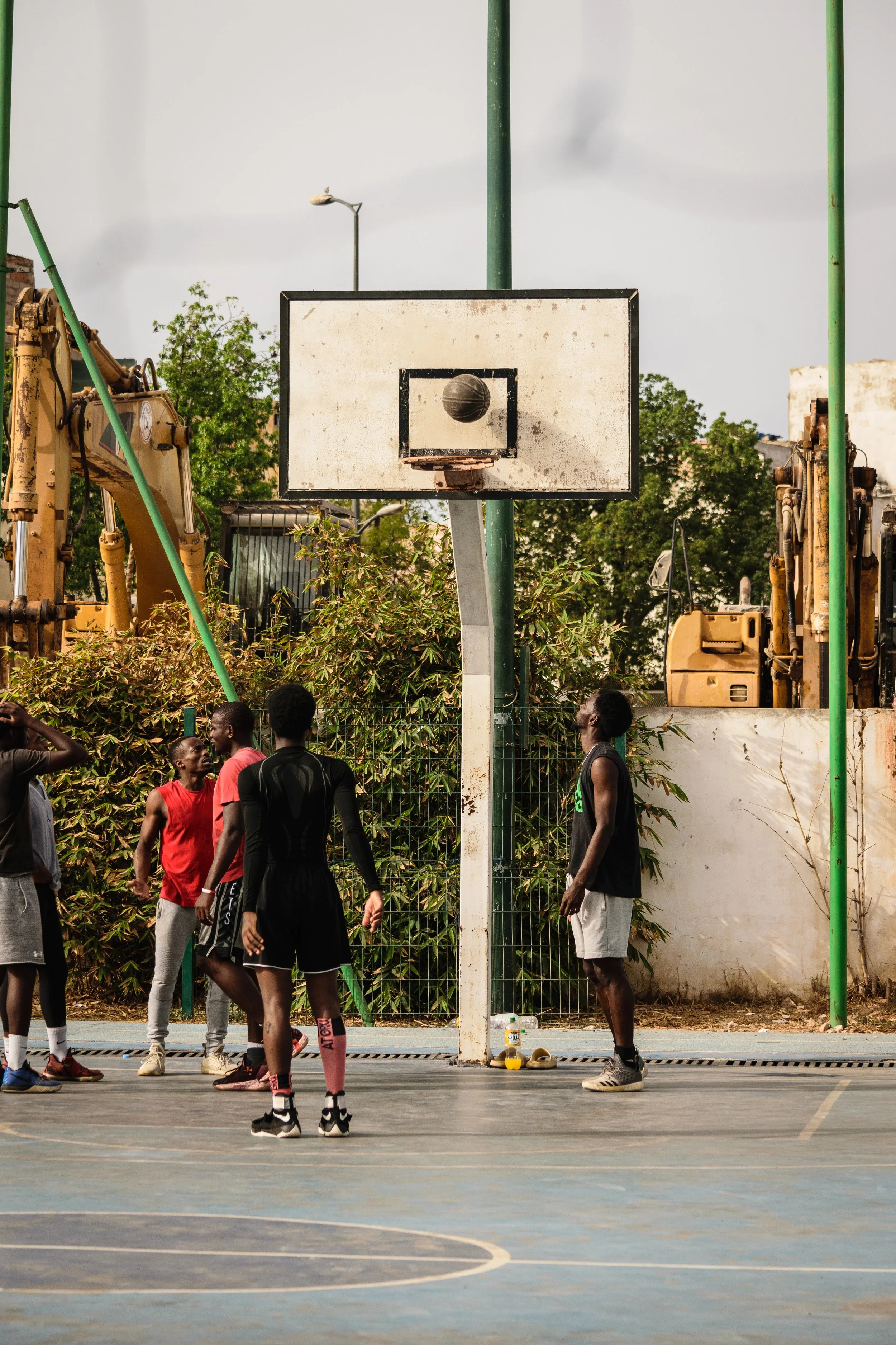 Group of young men playing basketball on an outdoor court; basketball hoop, construction equipment, and trees in background.