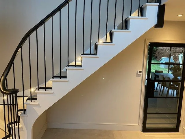 Interior view of a modern home showing a staircase with black railing and white steps, and a sliding glass door leading to an outdoor patio with a table and chairs.