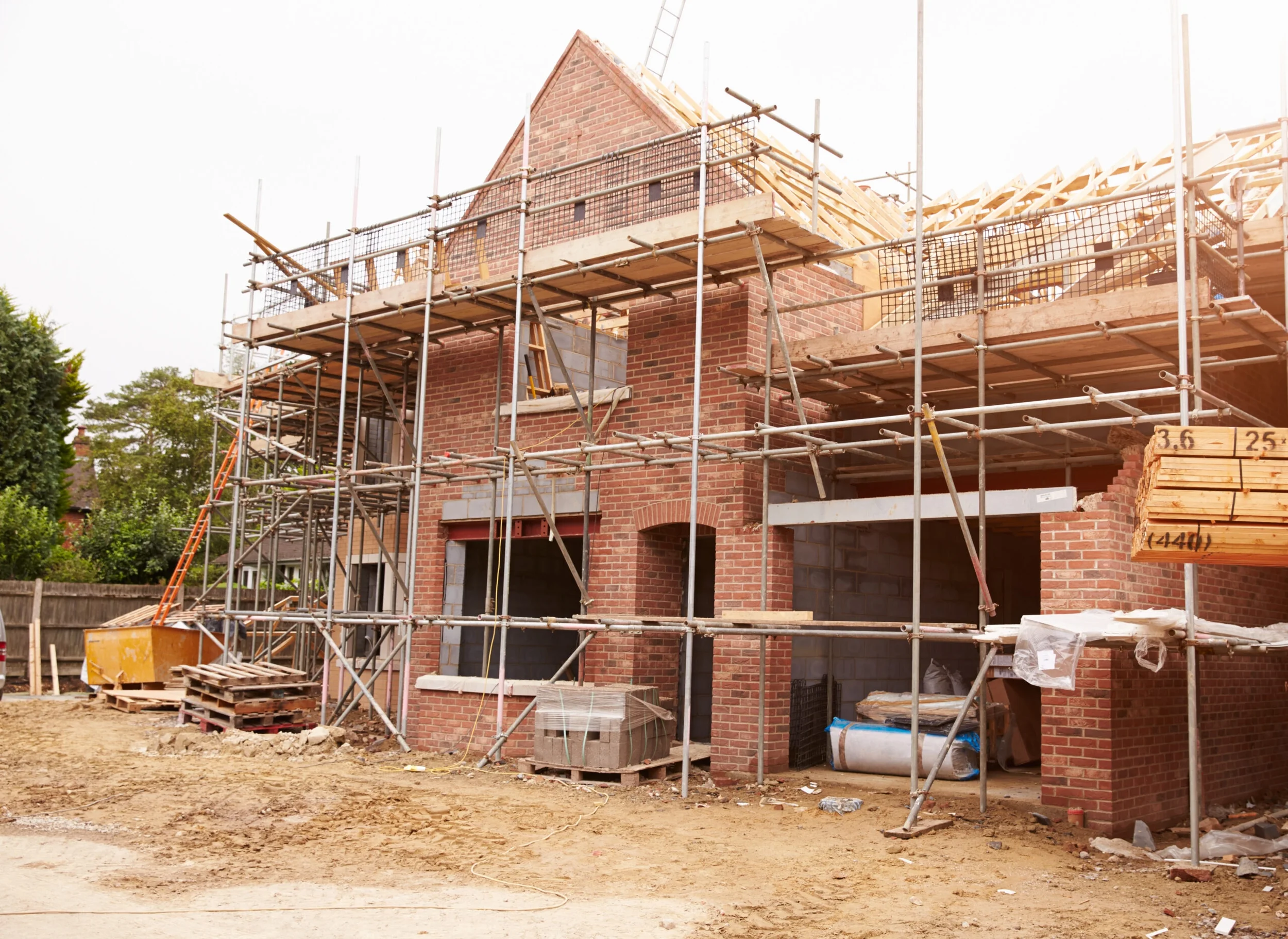 Construction site of a brick house with scaffolding around it. The house has a peaked roof, and construction materials and tools are scattered around the area.