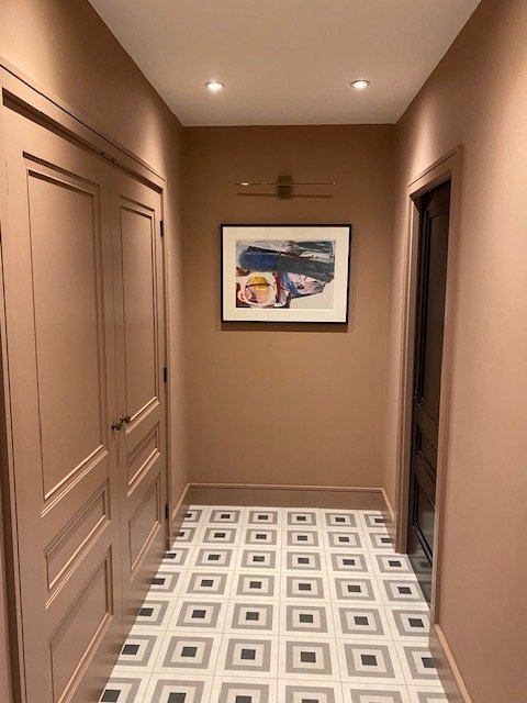 A narrow hallway with beige walls and baseboards, two recessed ceiling lights, a framed colorful abstract painting on the back wall, and a patterned tile floor with white, black, and gray geometric designs.